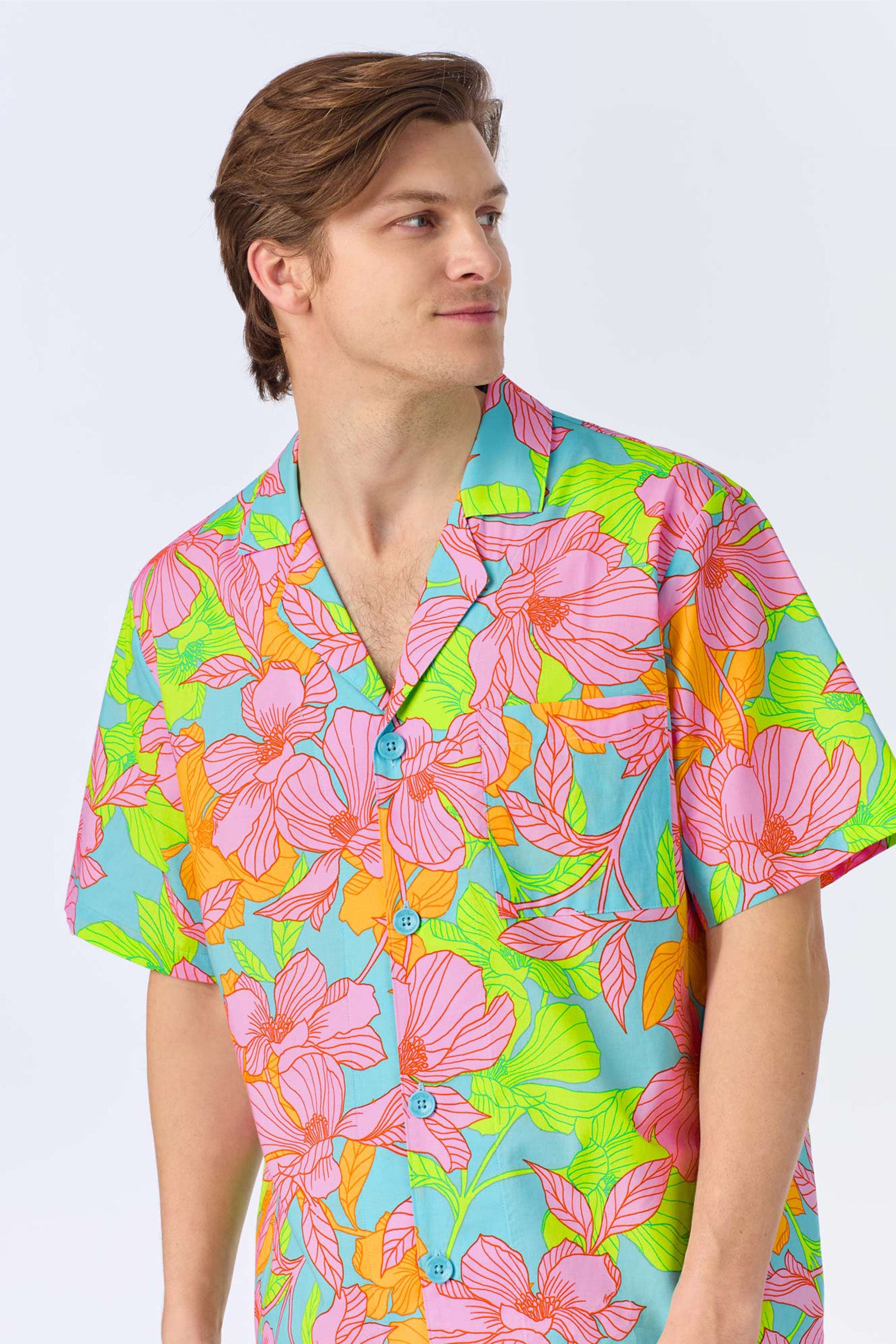 A man with light brown hair wearing the Mens Boxer PJ Set - Linear Floral Poplin, featuring a short-sleeve button-up shirt with a vibrant pink, green, and orange floral pattern on light blue, looks to his left.