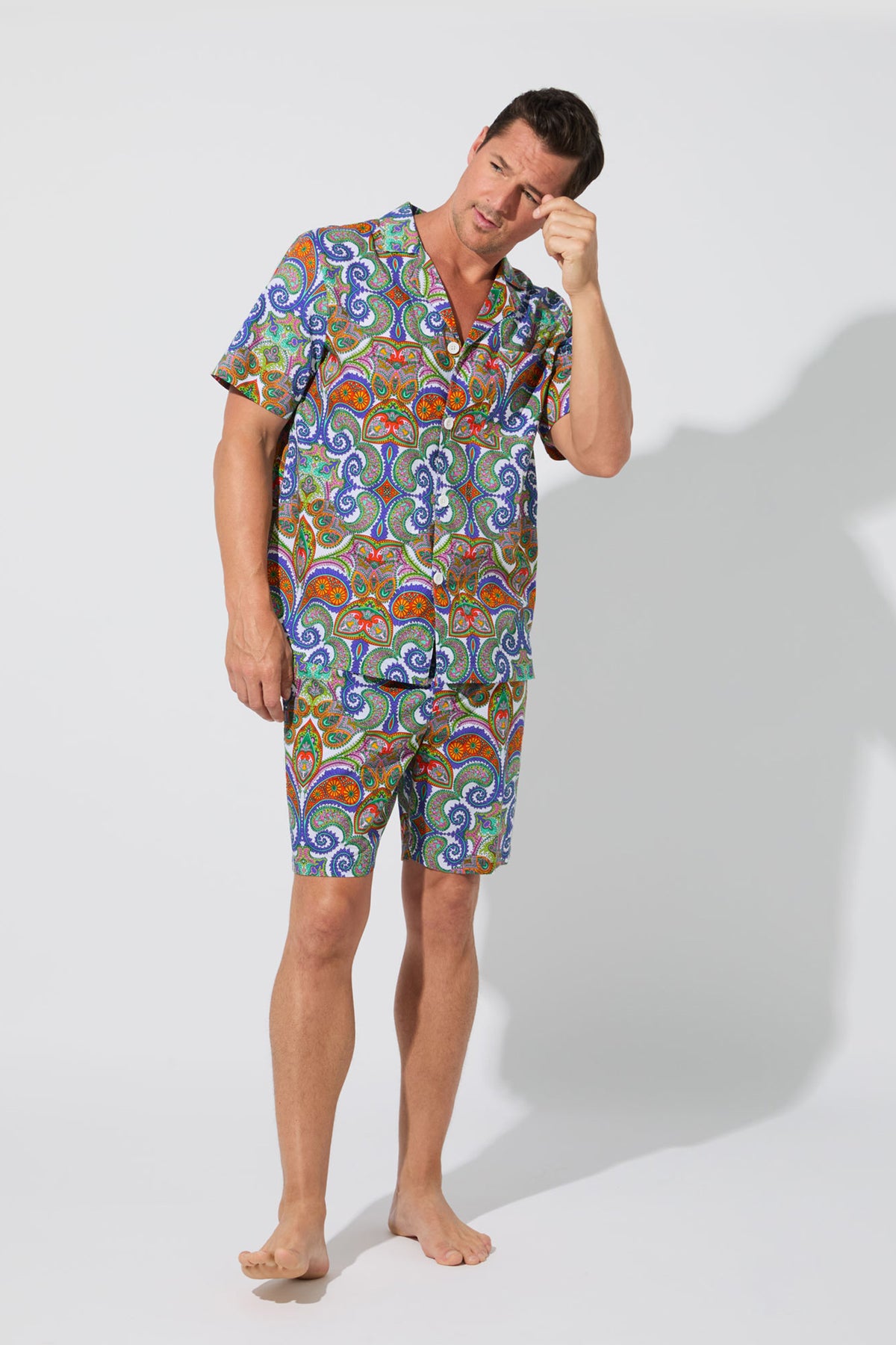 A man in the Mens Boxer PJ Set - Paisley, featuring colorful paisley-patterned short sleeves and matching shorts, stands barefoot against a plain white background, touching his forehead thoughtfully.
