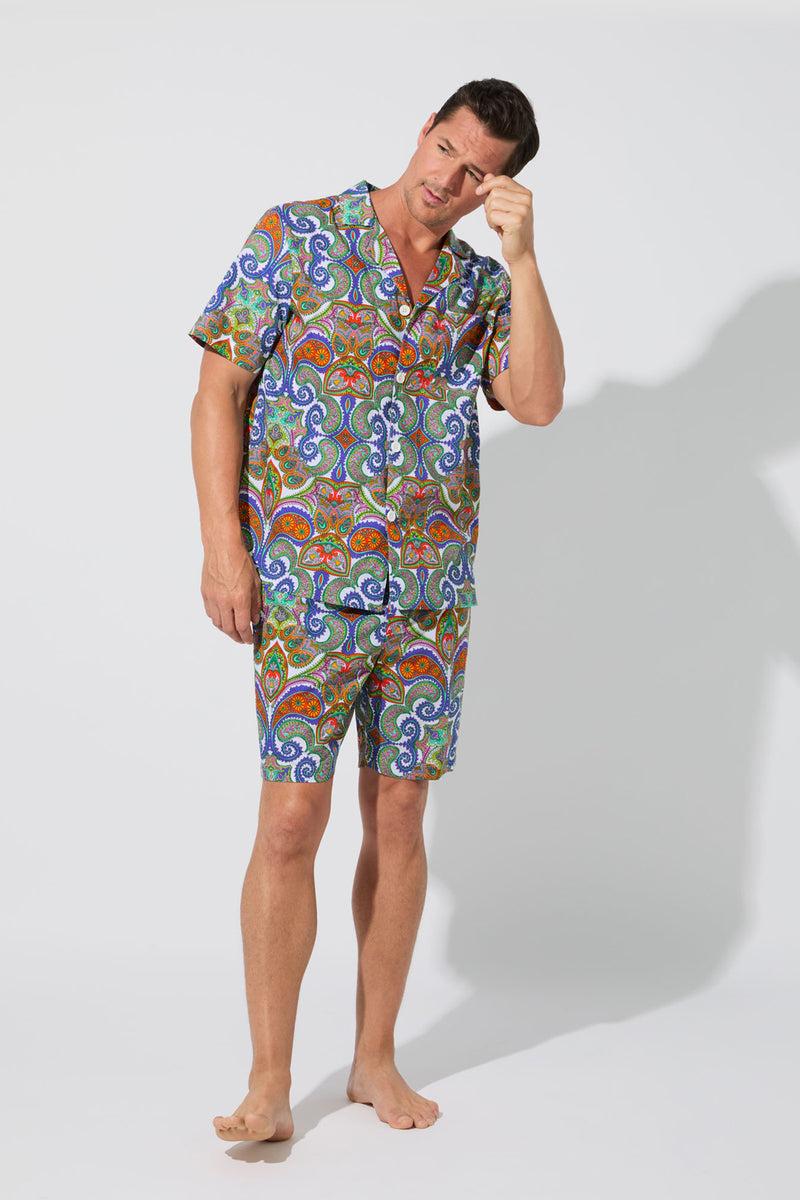 A man in the Mens Boxer PJ Set - Paisley, featuring colorful paisley-patterned short sleeves and matching shorts, stands barefoot against a plain white background, touching his forehead thoughtfully.