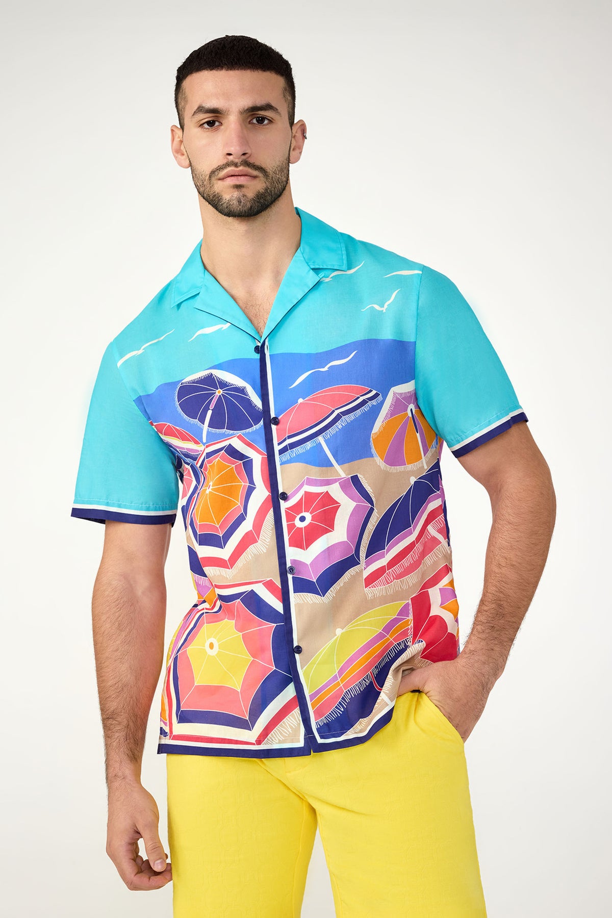 A man stands against a plain light background, looking serious, dressed in the Josue Short-Sleeve Shirt in Multi and yellow pants.