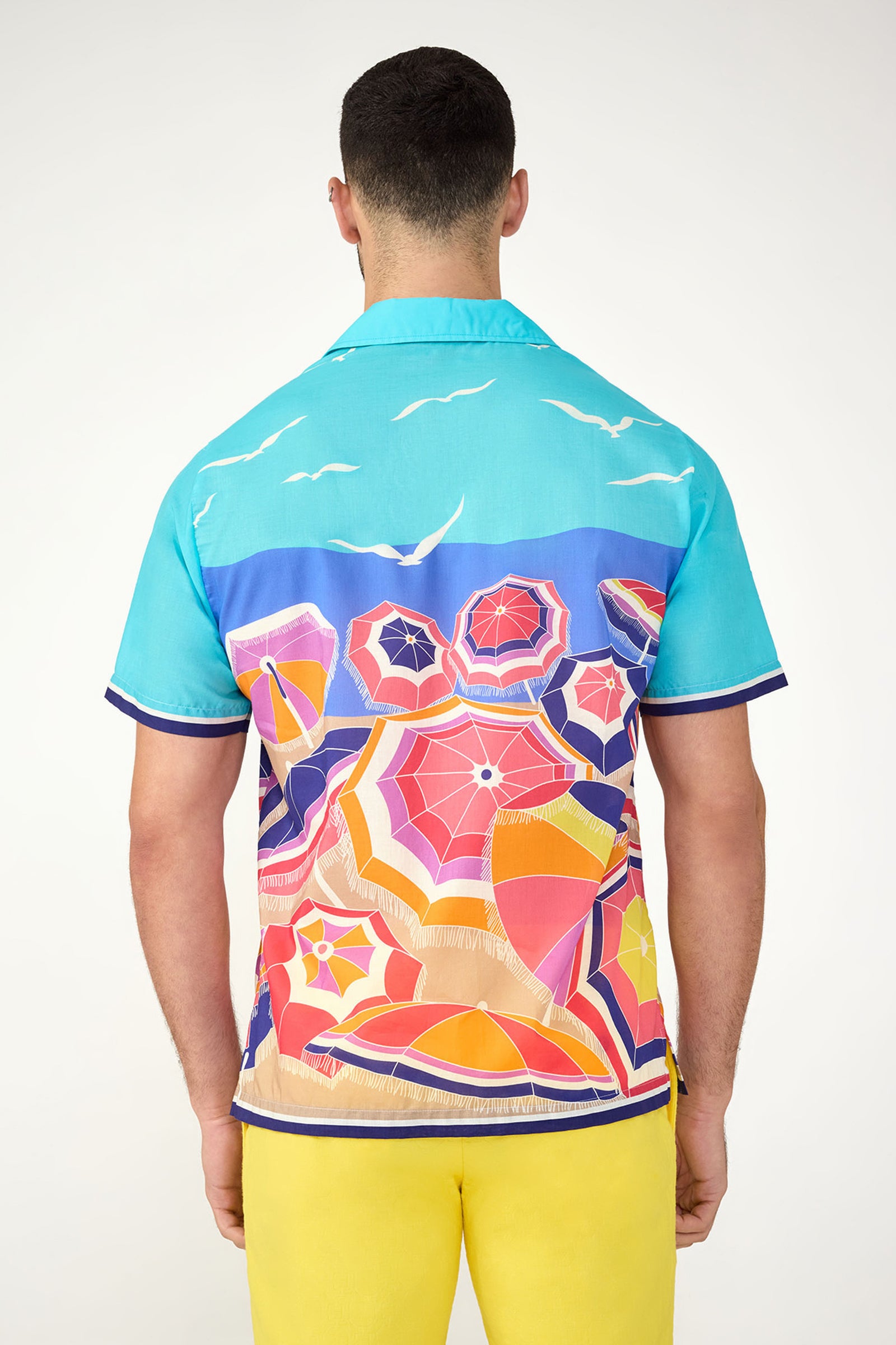 A man stands with his back visible, wearing the Josue Short-Sleeve Shirt in bright blue with beach umbrella, sand, and seagull motifs. He pairs it with yellow shorts against a plain light background.