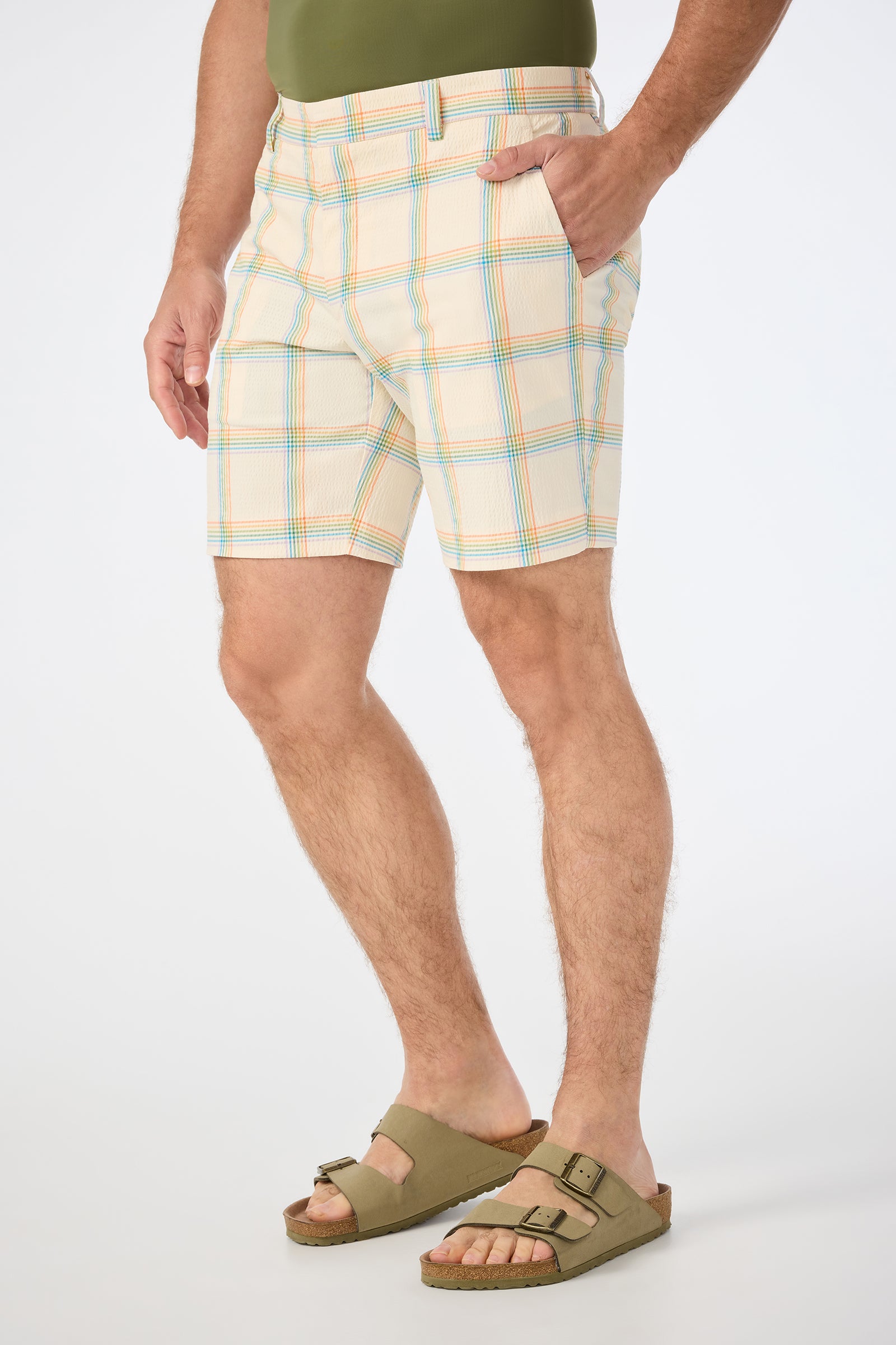 A person stands with one hand in their pocket against a plain background, wearing Travis Short - Pride Plaid, an olive green shirt, and matching Birkenstock sandals.