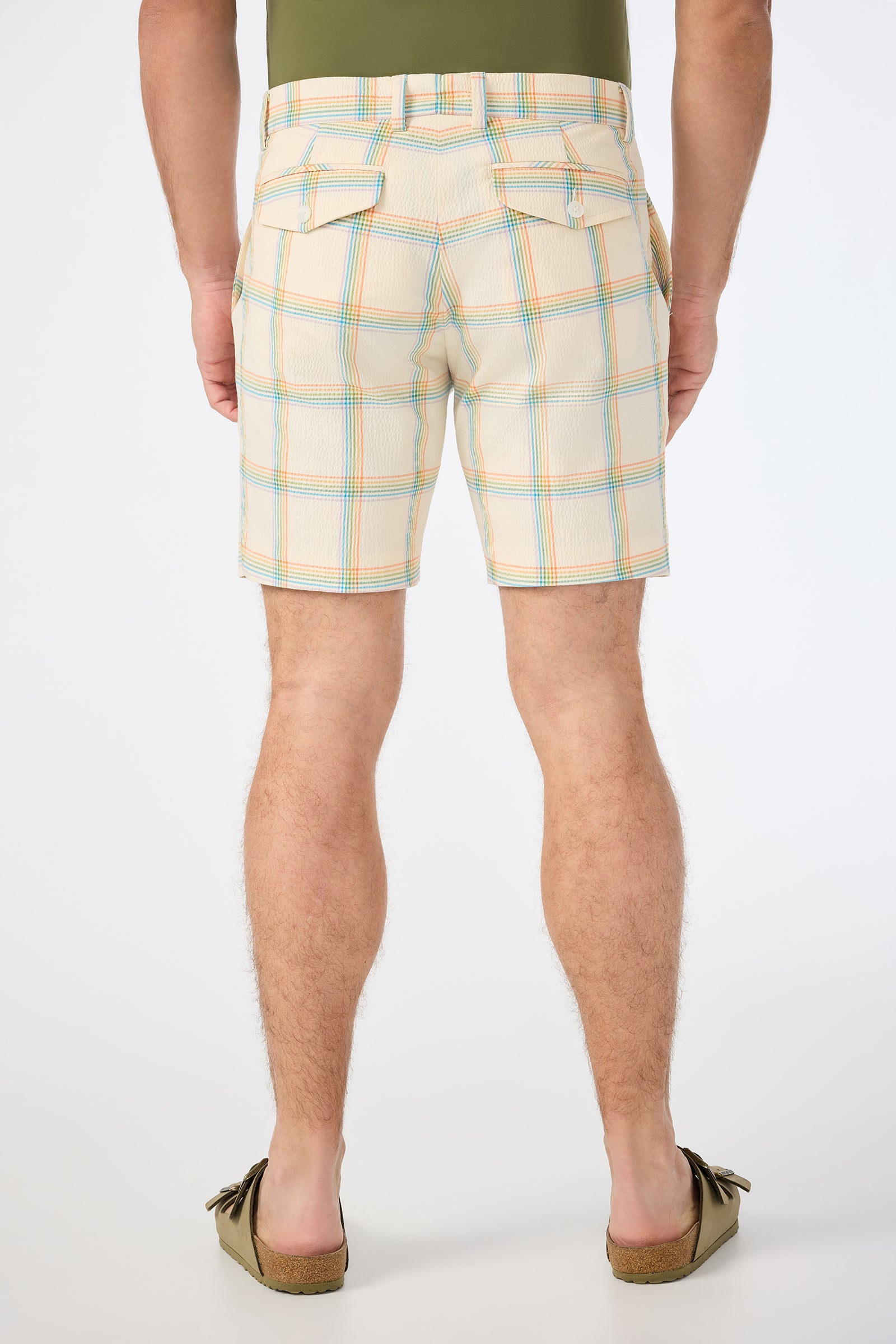 A person stands facing away, wearing the Travis Short - Pride Plaid (beige with blue and orange stripes), a green tucked-in shirt, and brown moccasins against a plain white background.