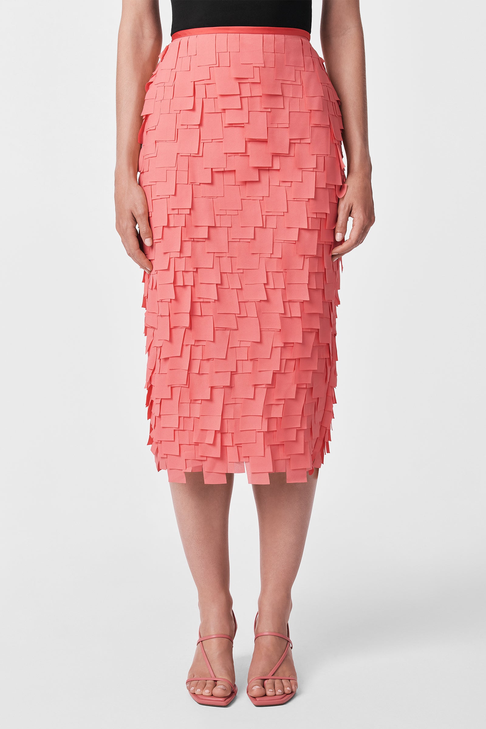 A person wears the Dita Midi Pencil Skirt - Flutter Chiffon in coral pink with layered square accents, a black top, and strappy pink heels, standing against a plain white background.