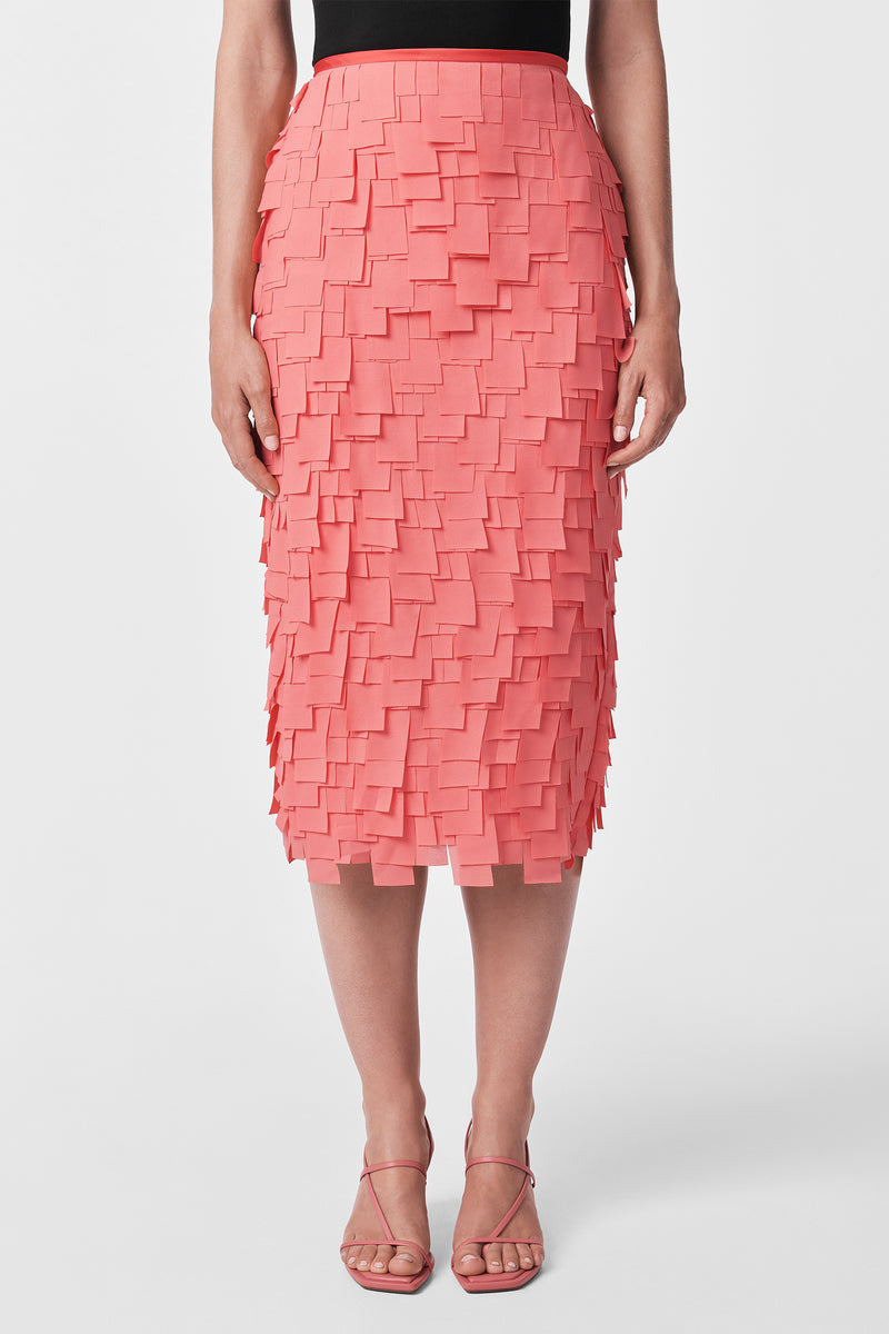 A person wears the Dita Midi Pencil Skirt - Flutter Chiffon in coral pink with layered square accents, a black top, and strappy pink heels, standing against a plain white background.