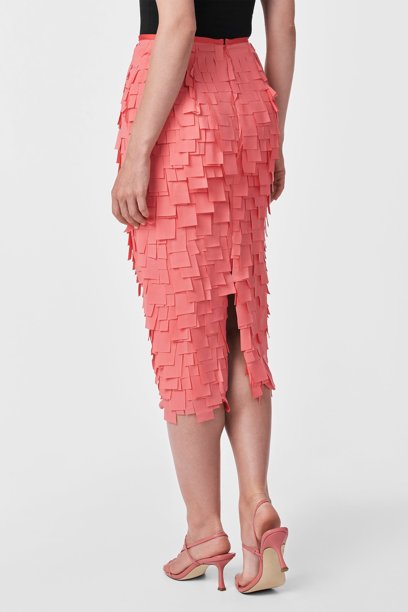 A person wears the Dita Midi Pencil Skirt - Flutter Chiffon in coral pink, featuring layered square chiffon pieces, paired with matching open-toe heels. The photo is taken from the back against a plain white background.