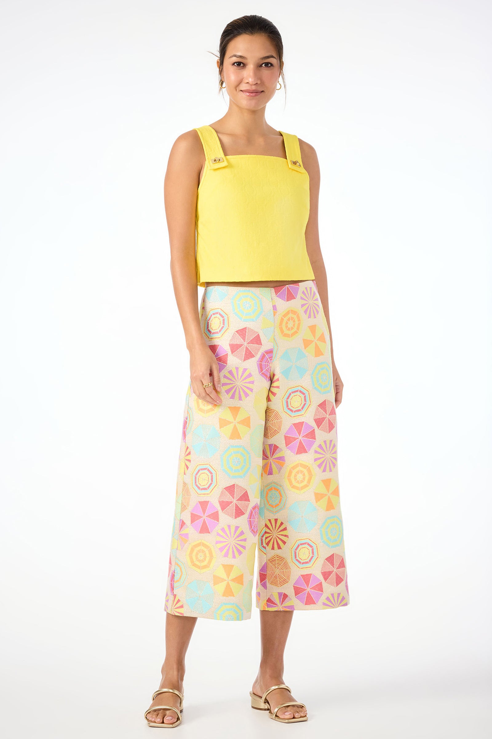 A woman smiles in Diona Crop Pants - Parapluie Jacquard, featuring a colorful pastel geometric print. She pairs them with a yellow sleeveless top and flat sandals, her hair tied back, posing against a plain white background.