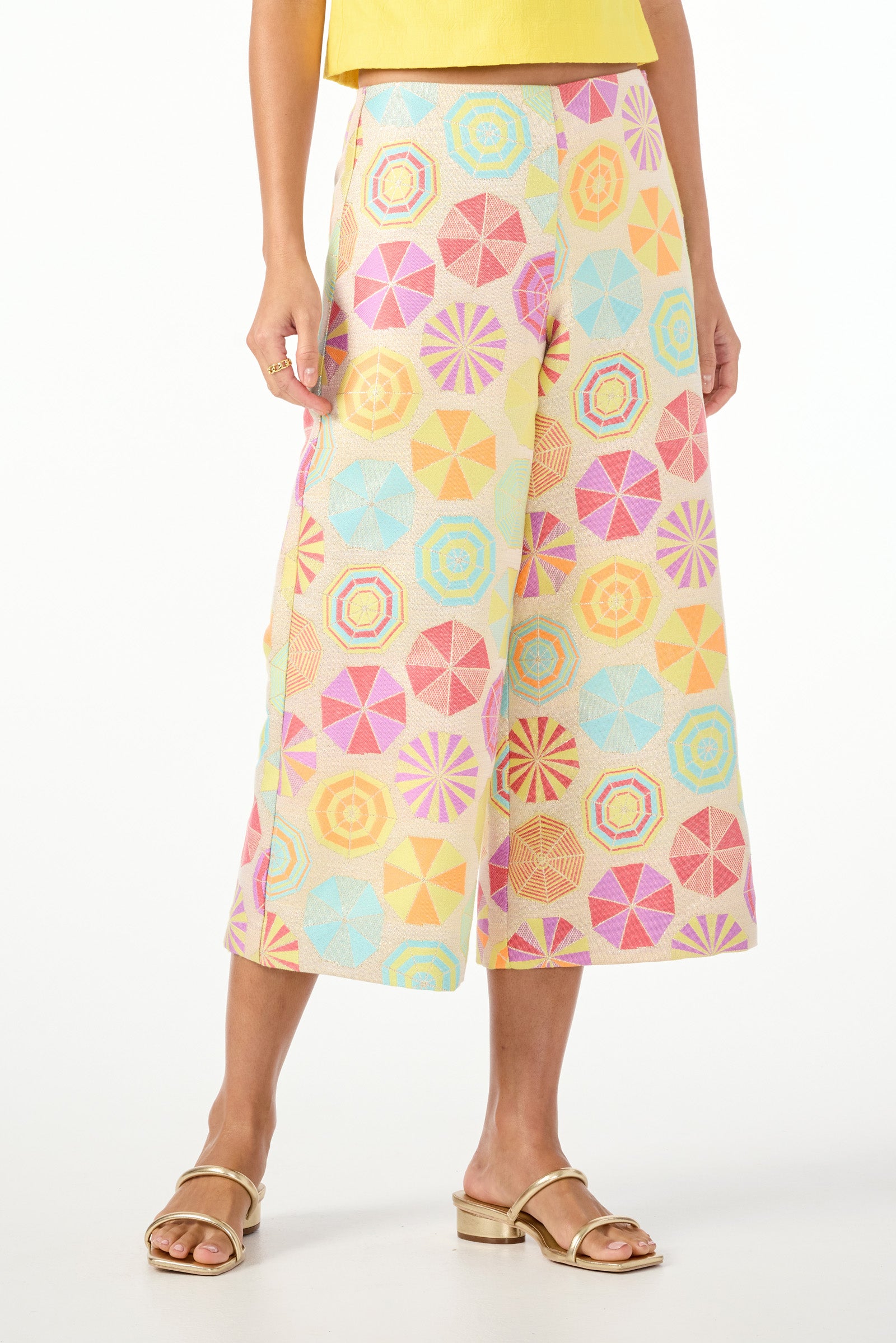 A person wears Diona Crop Pants in Parapluie Jacquard, featuring a colorful geometric pattern in pastel pink, blue, yellow, and orange paired with gold sandals against a white background.