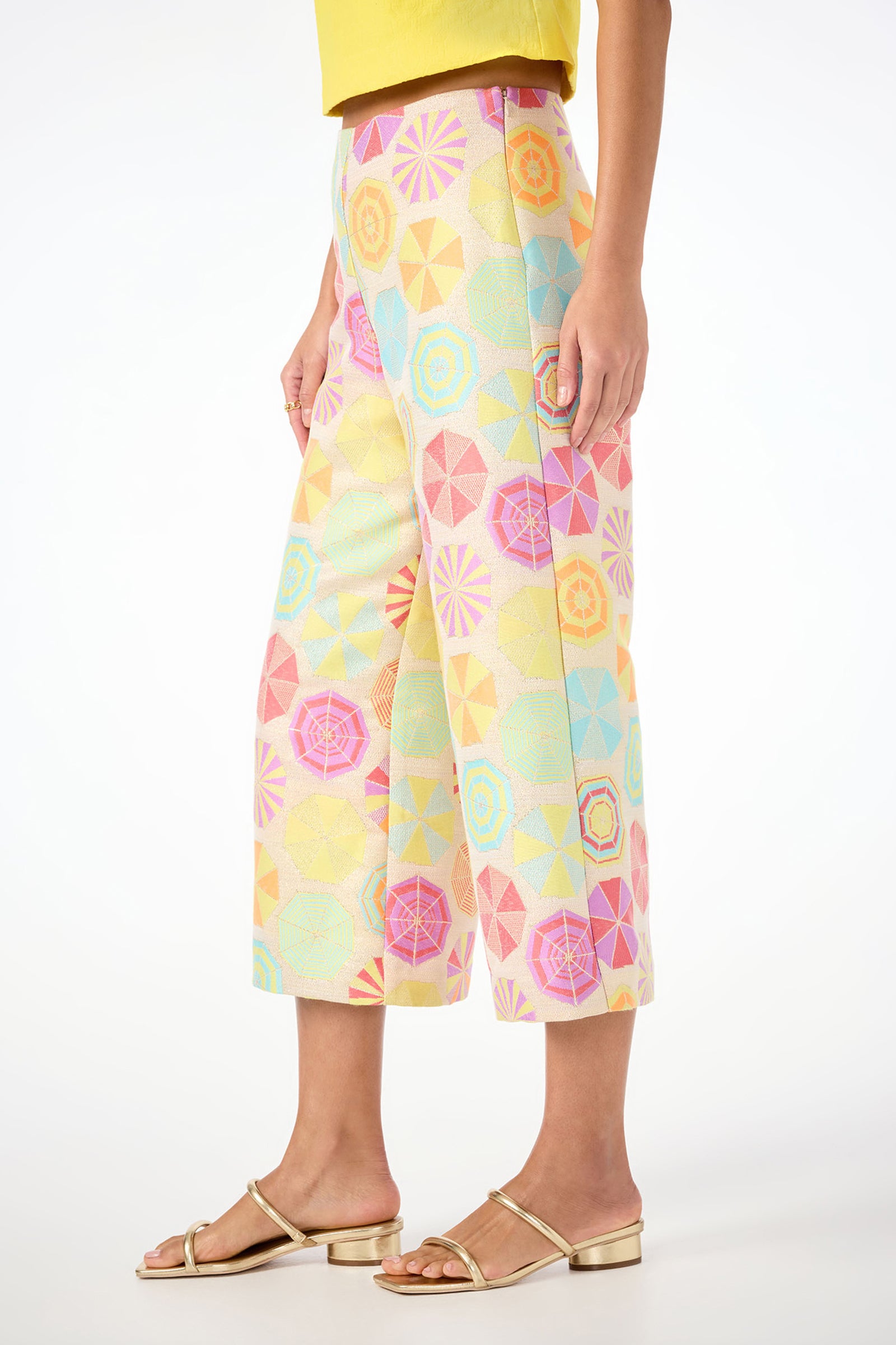 A person wearing the Diona Crop Pants - Parapluie Jacquard, featuring a pastel geometric pattern, along with a yellow top and gold sandals, stands against a white background.