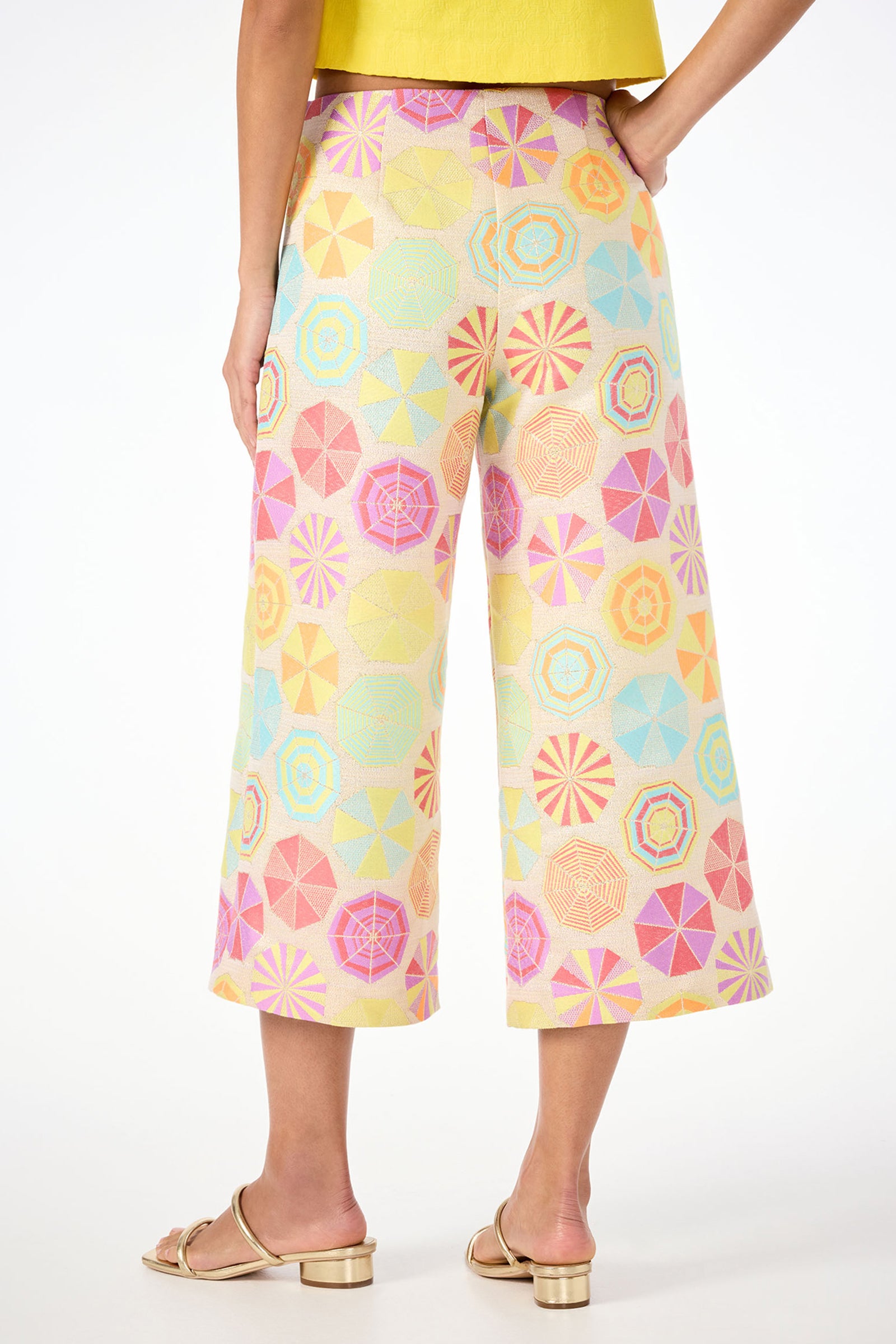 Shown from the back against a white background, a person wears Diona Crop Pants - Parapluie Jacquard with a colorful geometric umbrella pattern, gold sandals, and a yellow top.