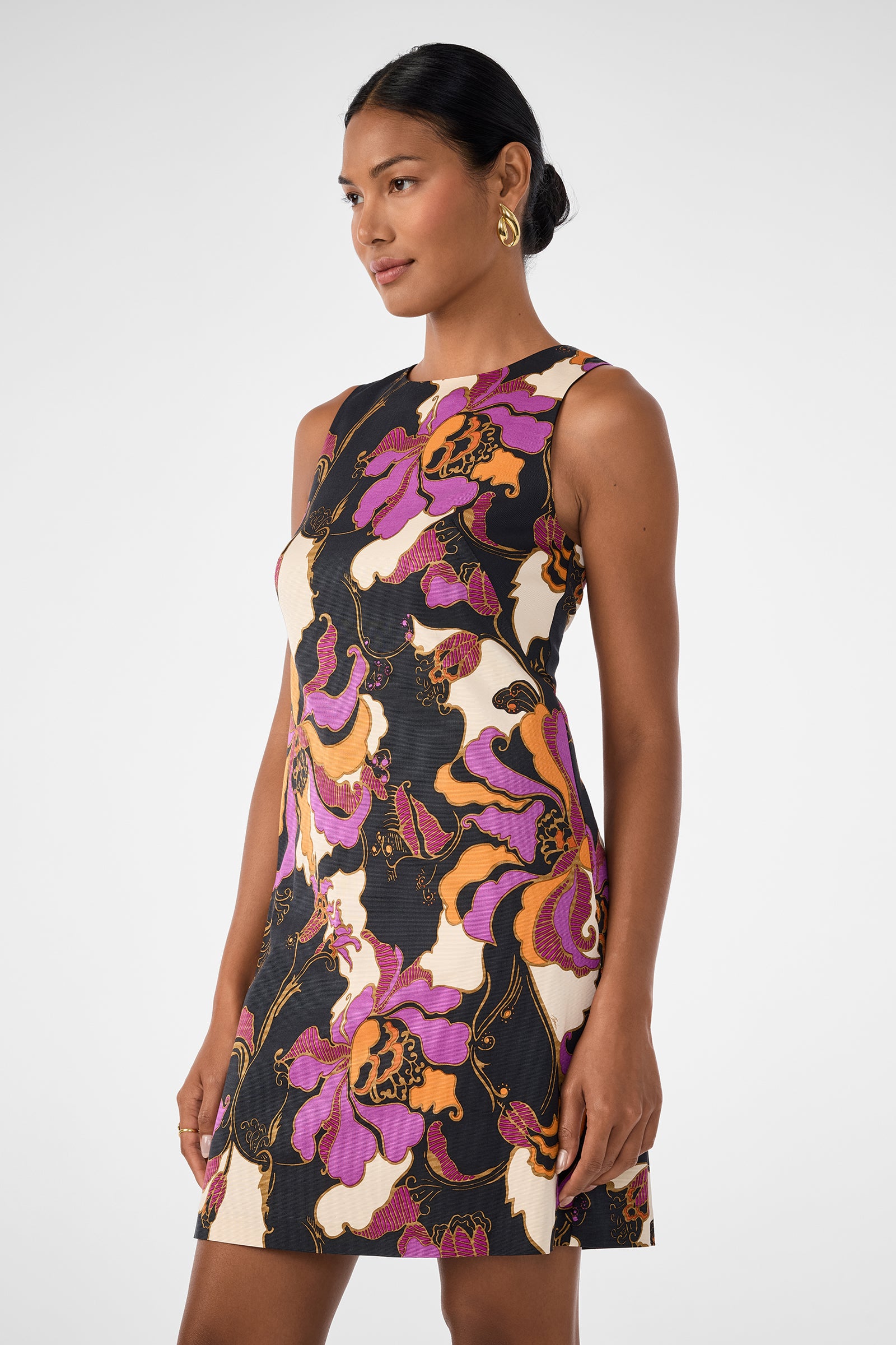 A woman models the Addy Dress—a sleeveless, fitted style with a bold floral print in purple, orange, black, and cream—her hair pulled back as she poses against a plain light background.