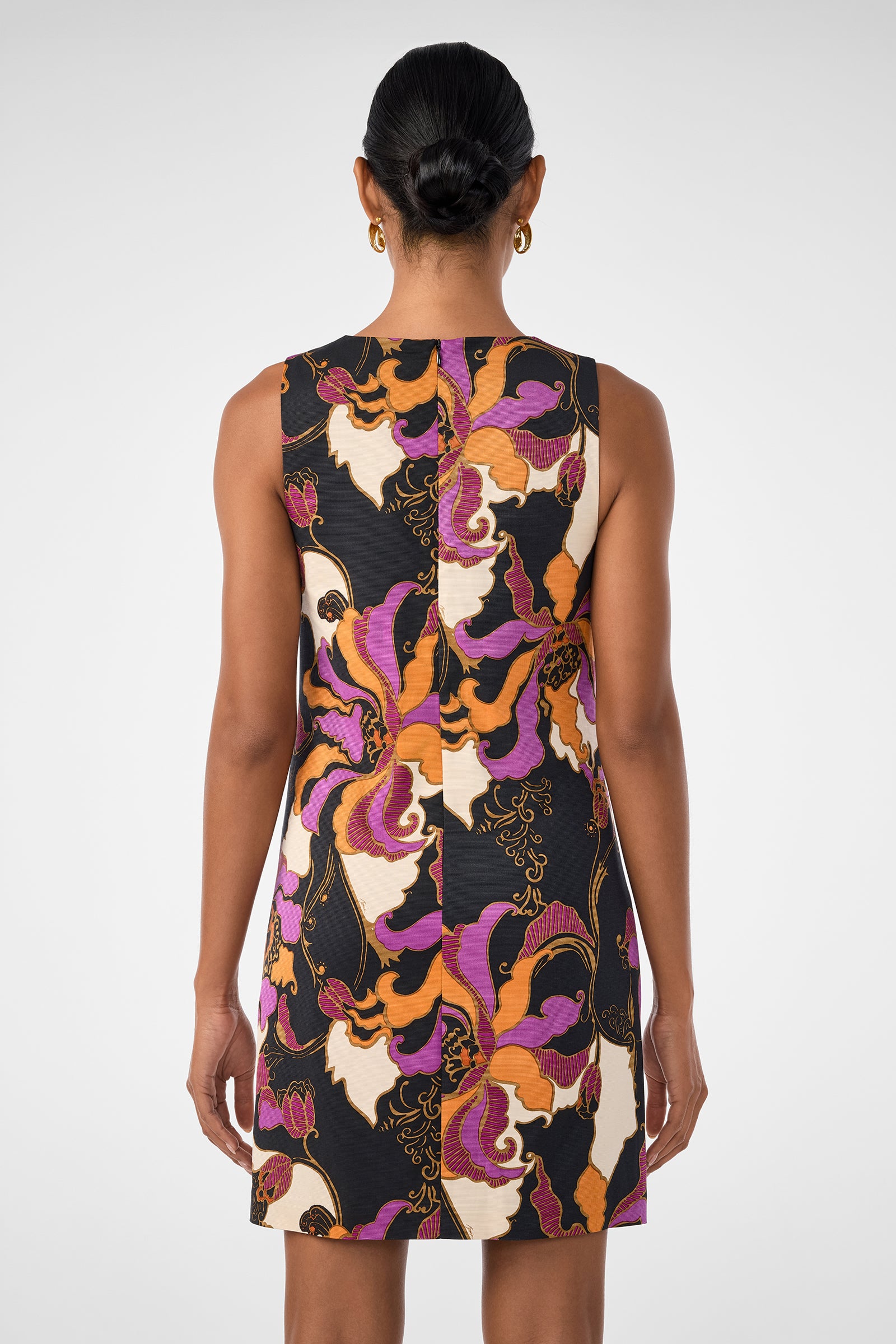 A woman with dark hair in a low bun is seen from behind wearing the Addy Dress, a sleeveless, above-knee style featuring a vibrant purple, orange, white, and black floral pattern. She accessorizes with gold hoop earrings.