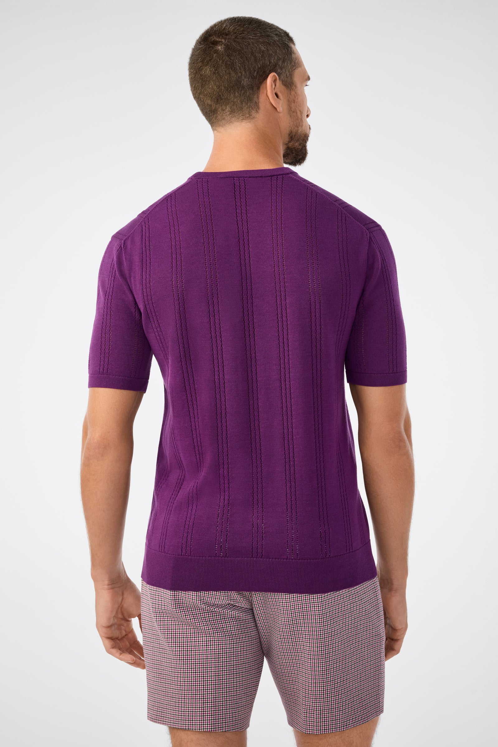 A man stands with his back to the camera in a Carlo Short-Sleeve Crewneck, purple knit, and plaid shorts against a plain light gray background.