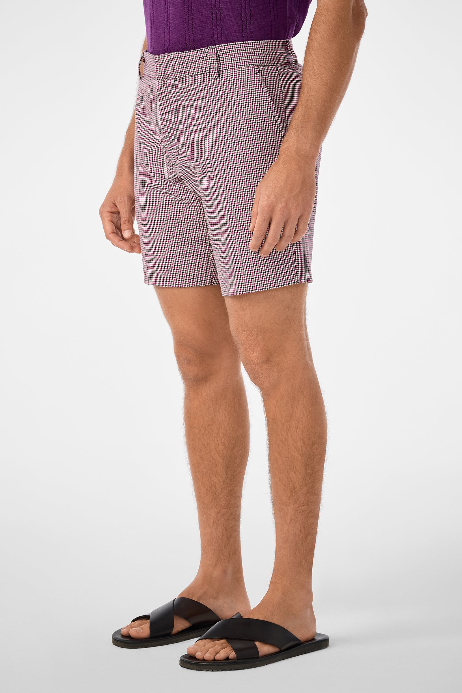 A man stands against a plain white background, angled slightly to the side with relaxed arms, wearing the Lawrence Short in red and white checks, a purple shirt, and black sandals.