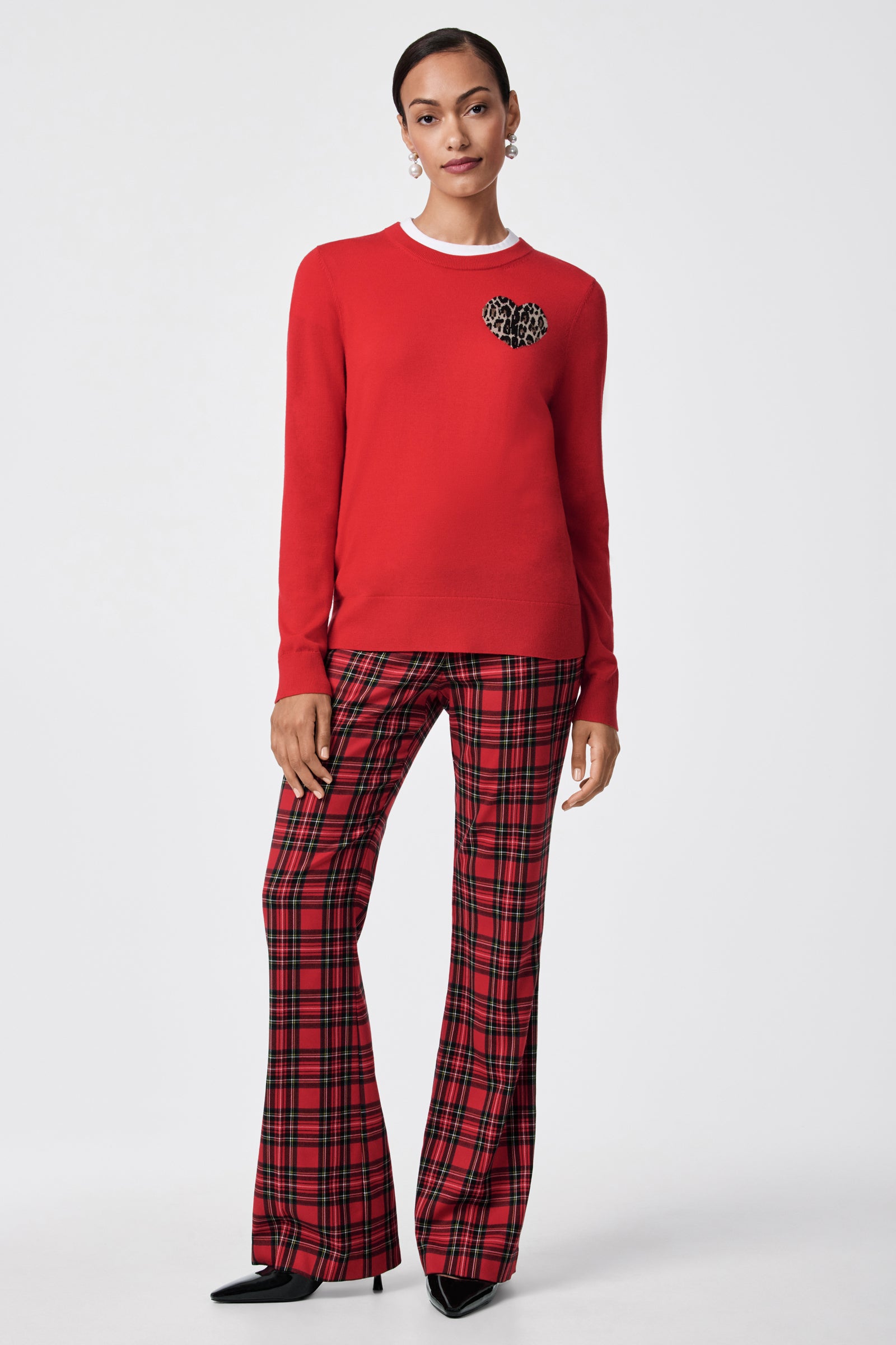 A woman stands against a plain background wearing the Jaguar Heart Crewneck Sweater, which is red with a small leopard print detail, paired with red plaid wide-leg pants, black pointed-toe shoes, and hoop earrings.