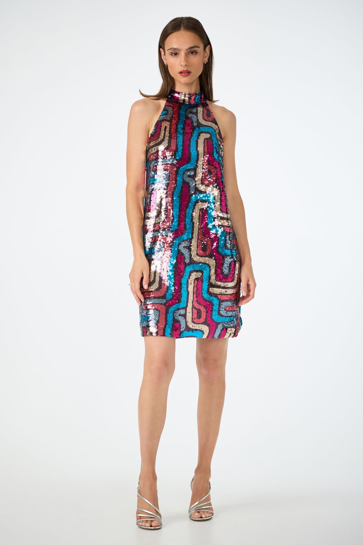 A woman faces forward in the Flamenco Sequin Mini Dress, a sleeveless, high-neck mini dress with colorful wavy sequins. She has straight brown hair, red lipstick, and wears silver strappy heels against a plain white background.