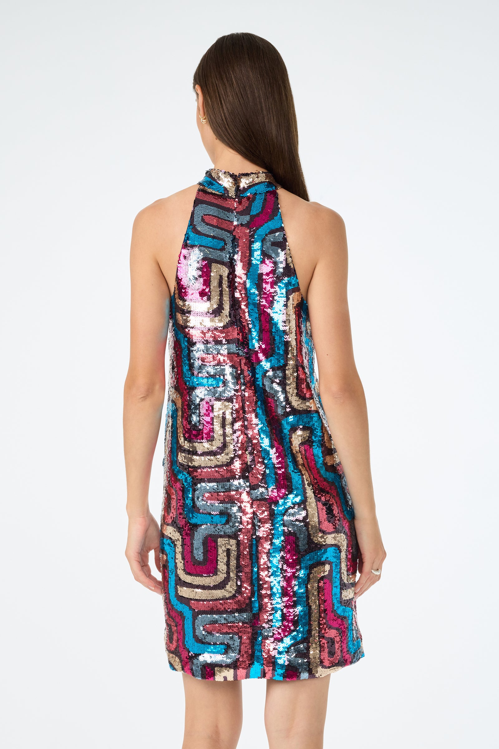 A woman faces away, wearing the Flamenco Sequin Mini Dress—a sleeveless, high-neck mini dress adorned with colorful sequins in a bold geometric pattern of pink, blue, black, and gold.