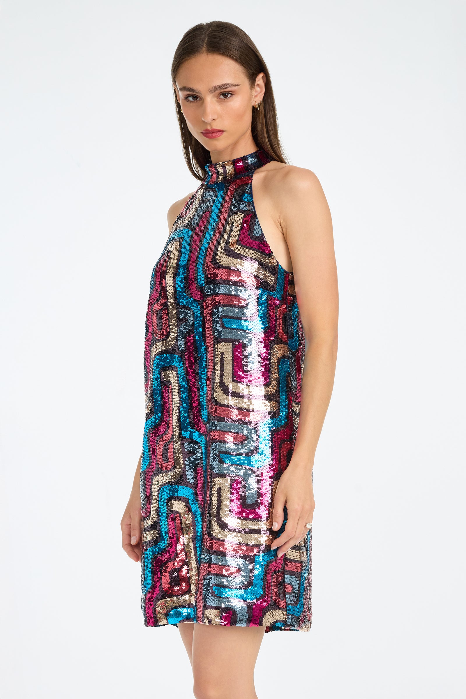 A woman stands against a white background wearing the Flamenco Sequin Mini Dress, a sleeveless, high-neck mini covered in colorful geometric sequins of blue, pink, gold, and black.