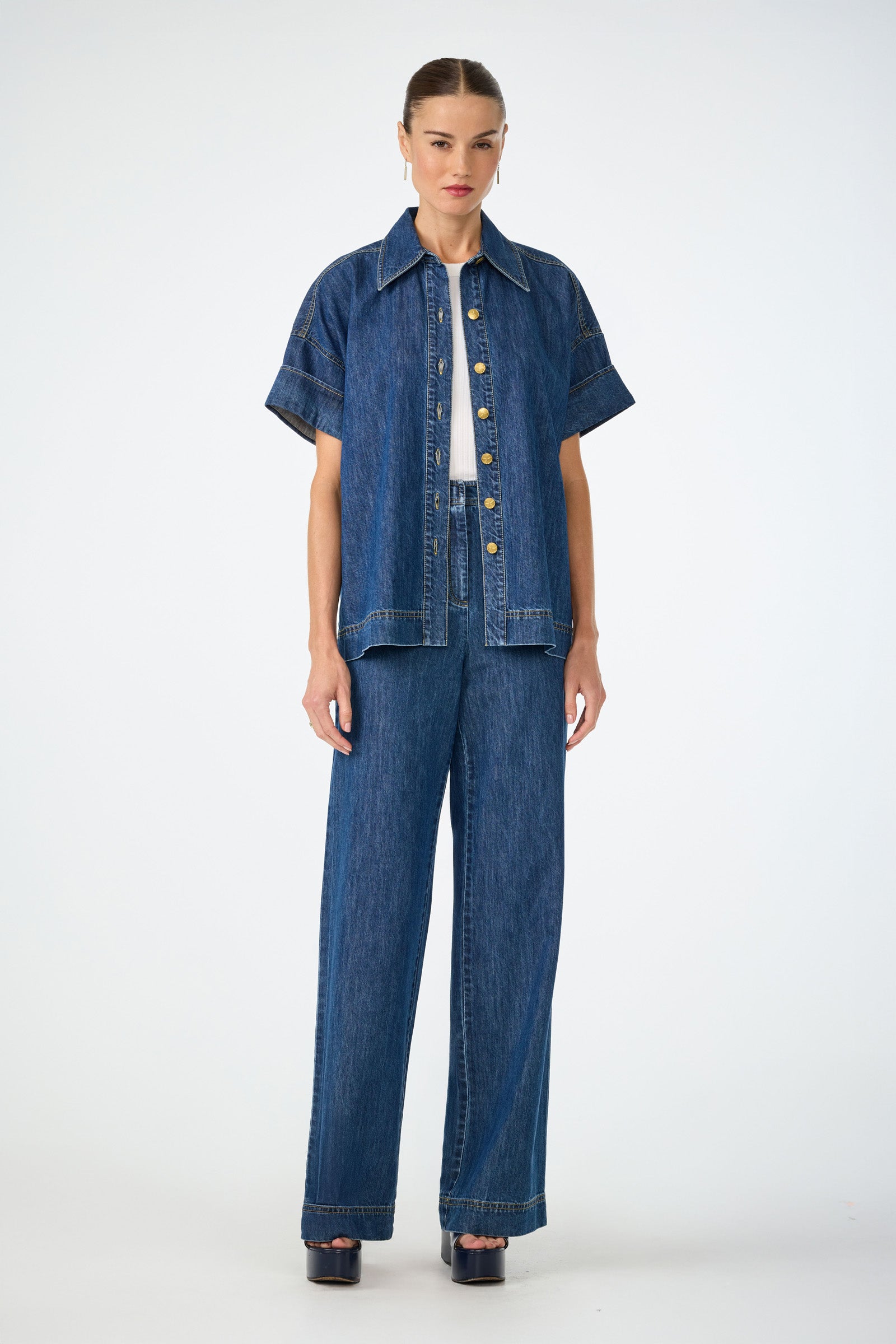 A woman faces forward wearing the Opponent 2 Short-Sleeve Top over a white top, paired with matching high-waisted wide-leg denim pants and black platform sandals, against a plain white background.