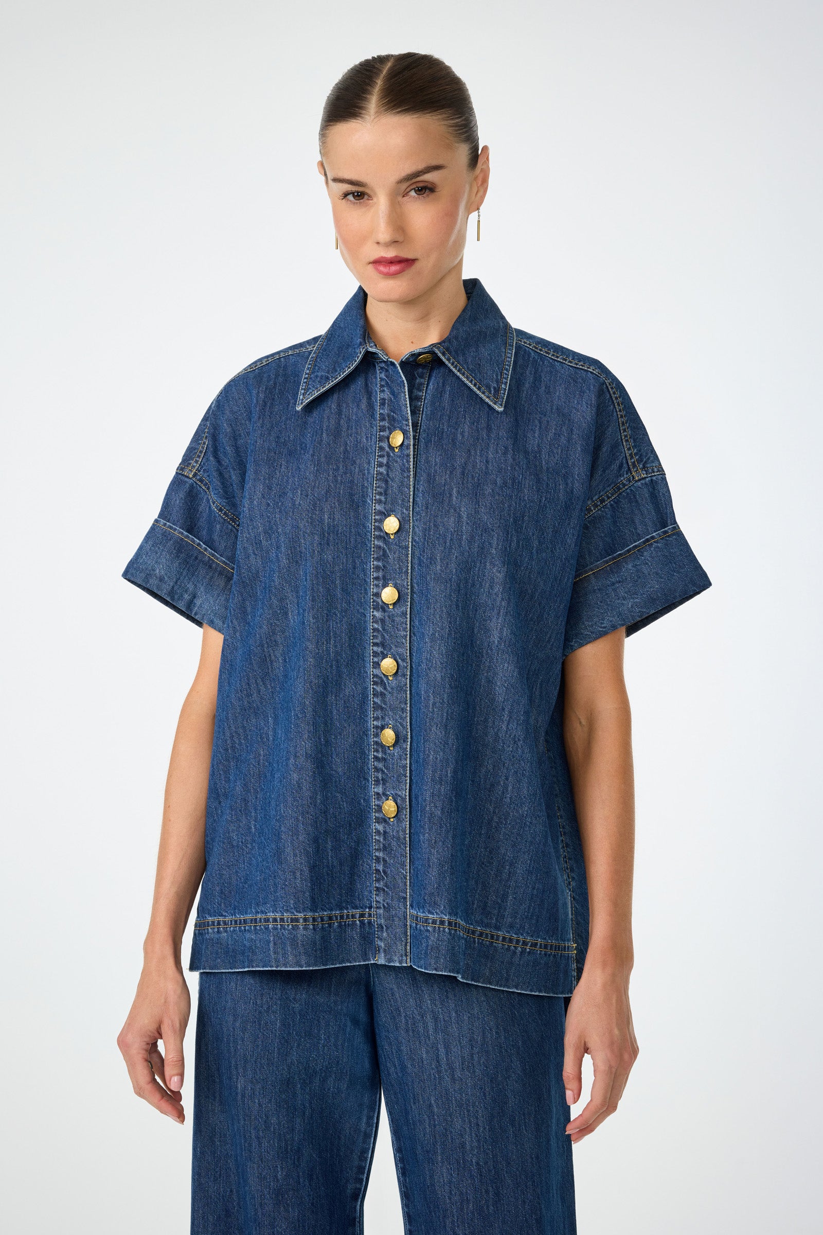 A woman with slicked-back hair and minimal makeup wears the indigo Opponent 2 Short-Sleeve Top, featuring large gold buttons, paired with matching wide-leg denim pants against a plain background. She accessorizes with earrings.