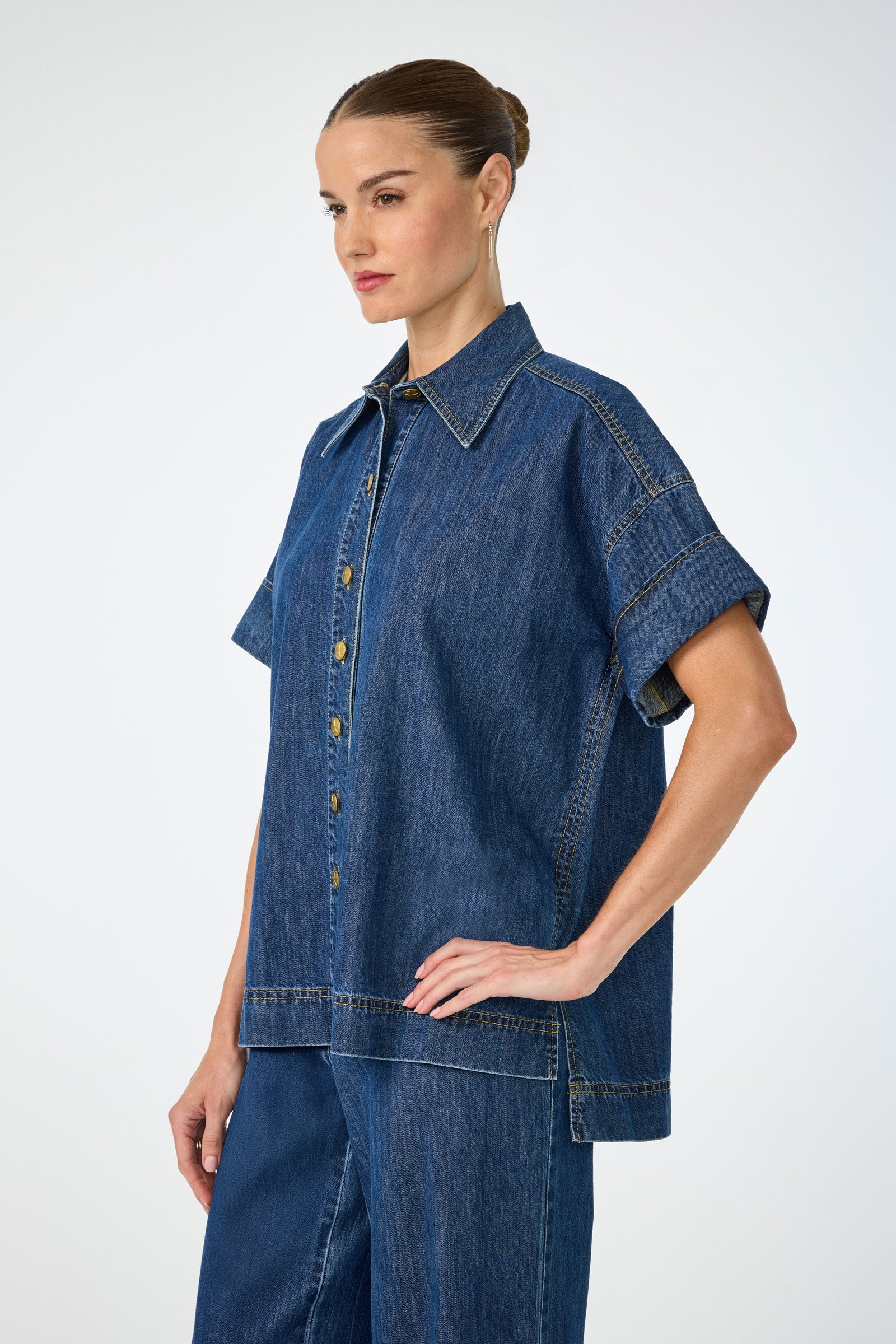 A woman with brown hair in a bun models the Opponent 2 Short-Sleeve Top and matching denim pants, standing in profile with one hand on her hip against a plain light background.