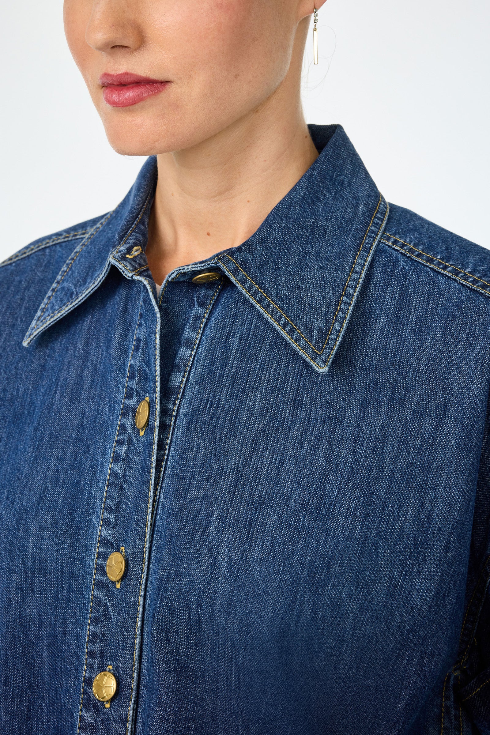A close-up of a woman wearing the Opponent 2 Short-Sleeve Top, a blue denim shirt with gold buttons and yellow stitching. Image highlights the collar, upper chest, gold earring, and her pink lipstick.