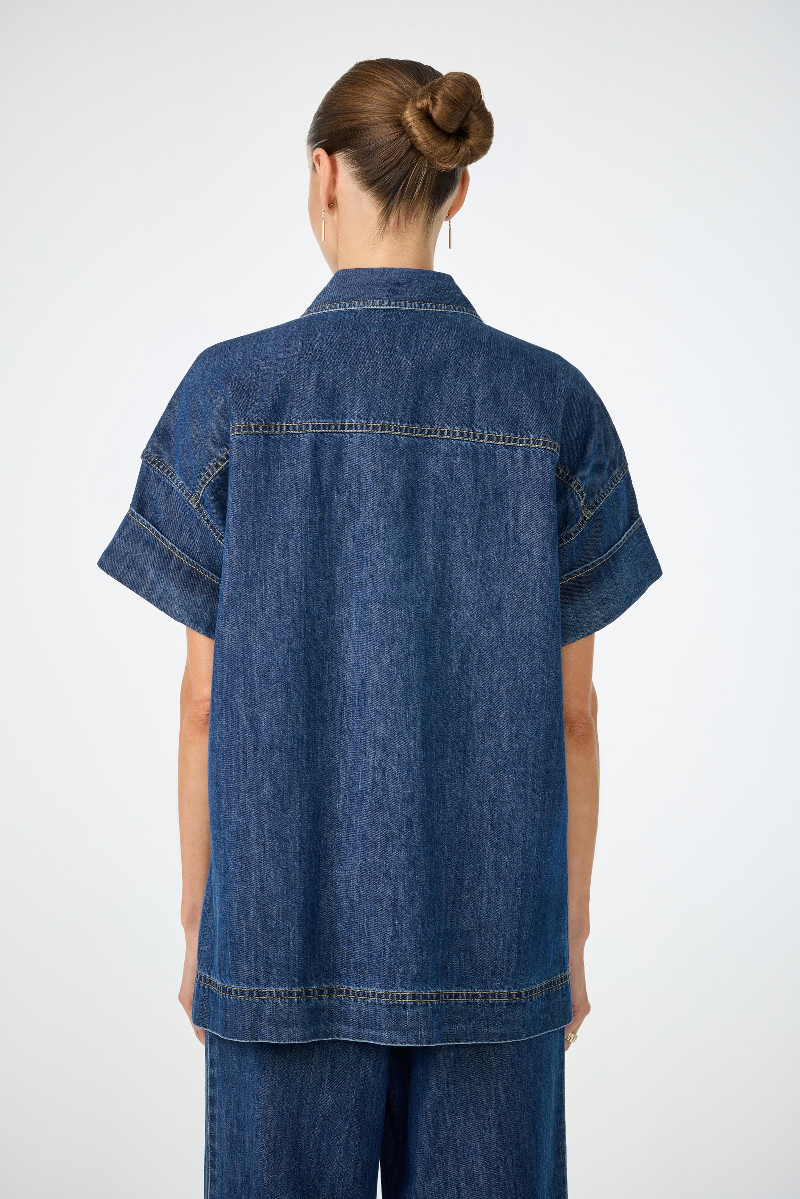 A woman with brown hair in a bun is shown from behind, wearing the Opponent 2 Short-Sleeve Top and matching denim pants against a plain light background.