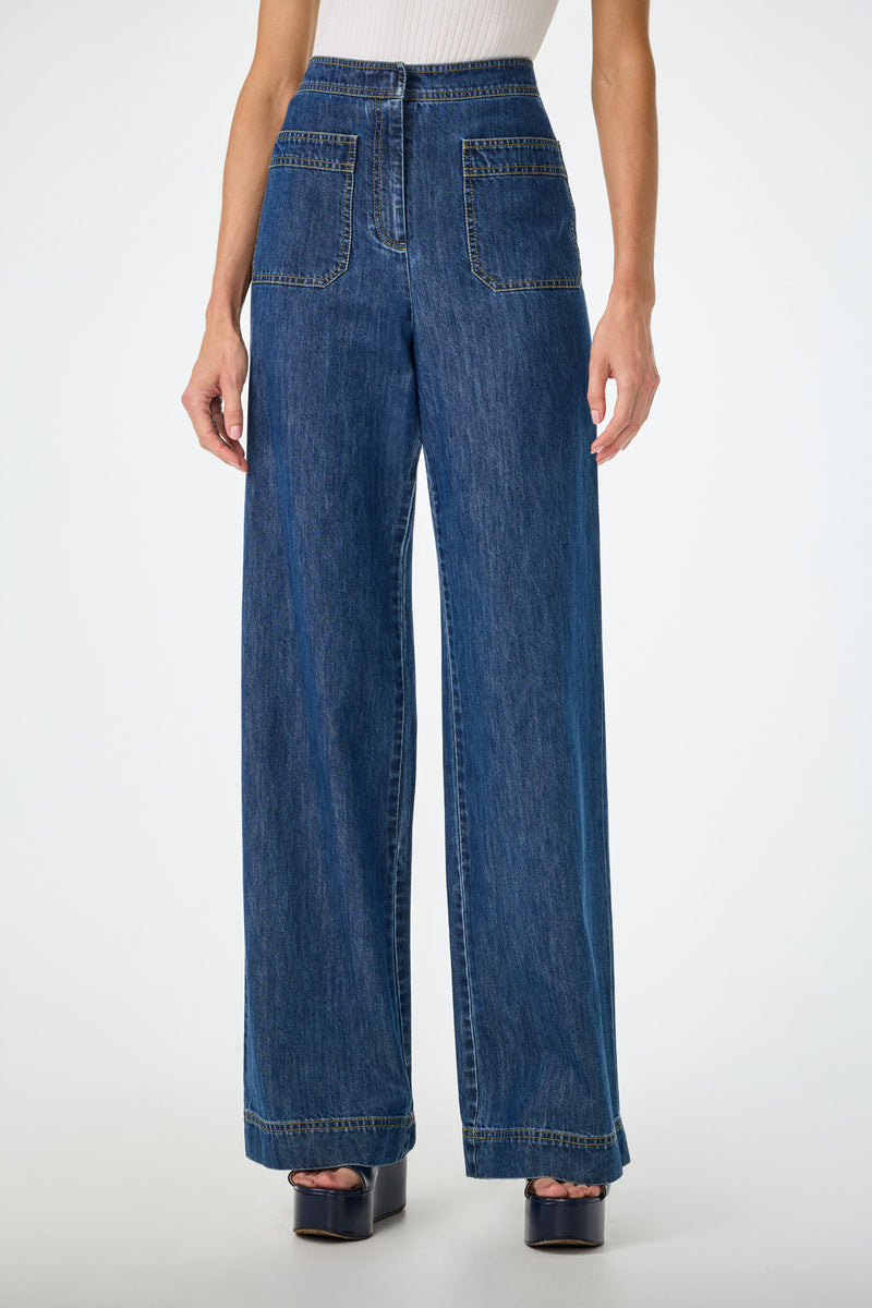 Someone wearing the Ferrer Wide Leg Jean in Indigo, featuring high-waisted, wide-leg styling and large front patch pockets, paired with a sleeveless white ribbed top and platform sandals against a plain white background.