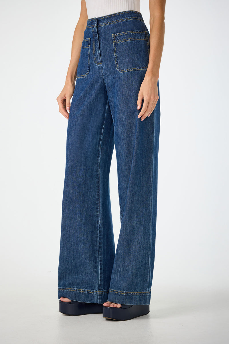A person wears Ferrer Wide Leg Jeans—high-waisted, wide-leg blue denim with front patch pockets—paired with platform sandals and a sleeveless white top, standing against a plain light background.