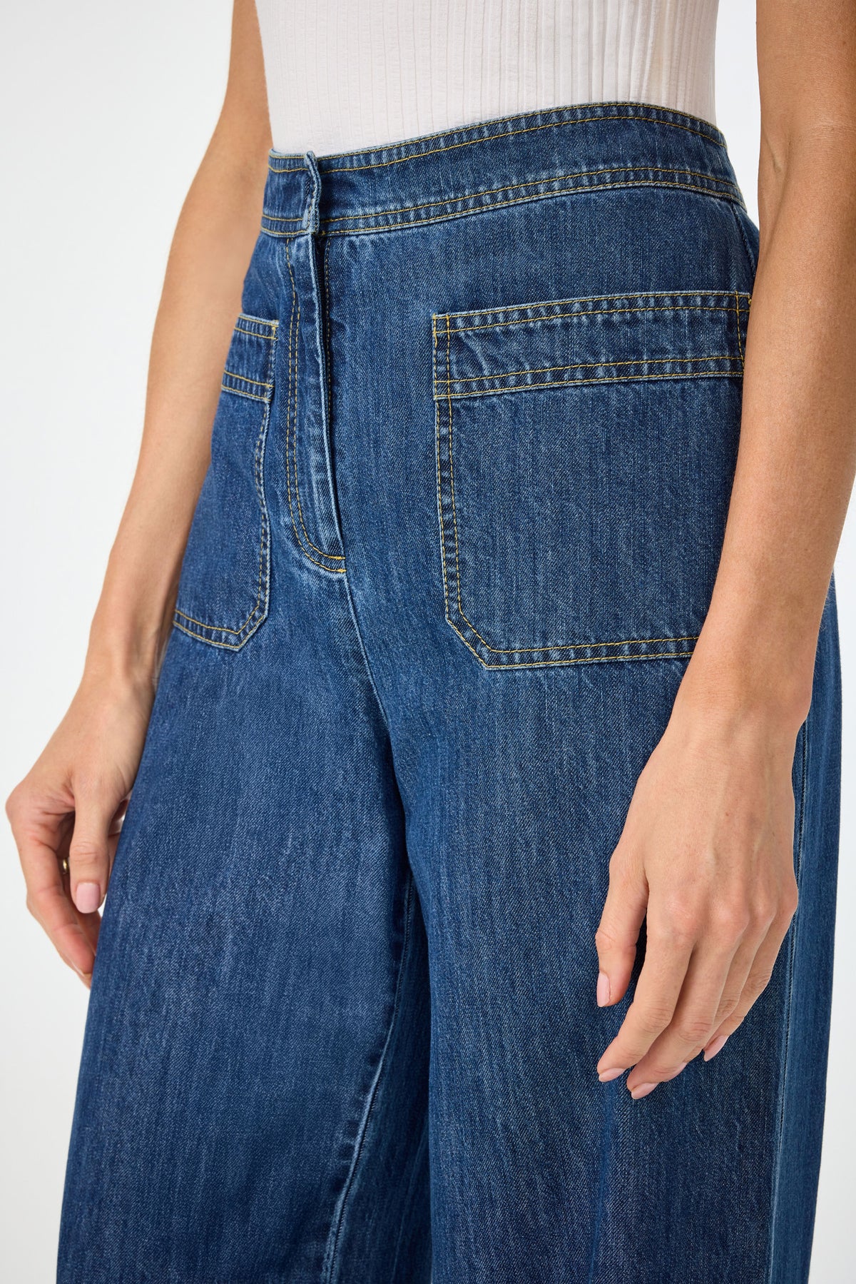 Wearing Ferrer Wide Leg Jean in high-waisted blue denim with large front pockets and visible stitching, paired with a white ribbed sleeveless top. Shown from the waist down.