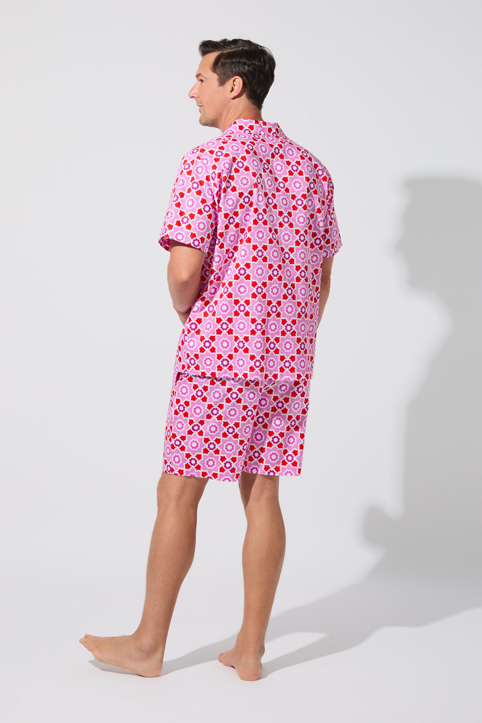 A barefoot man faces away, wearing the Mosiac Tile Mens S/S Boxer Set—matching pink shirt and shorts with a bold geometric circle and diamond pattern—against a plain, light-colored background.