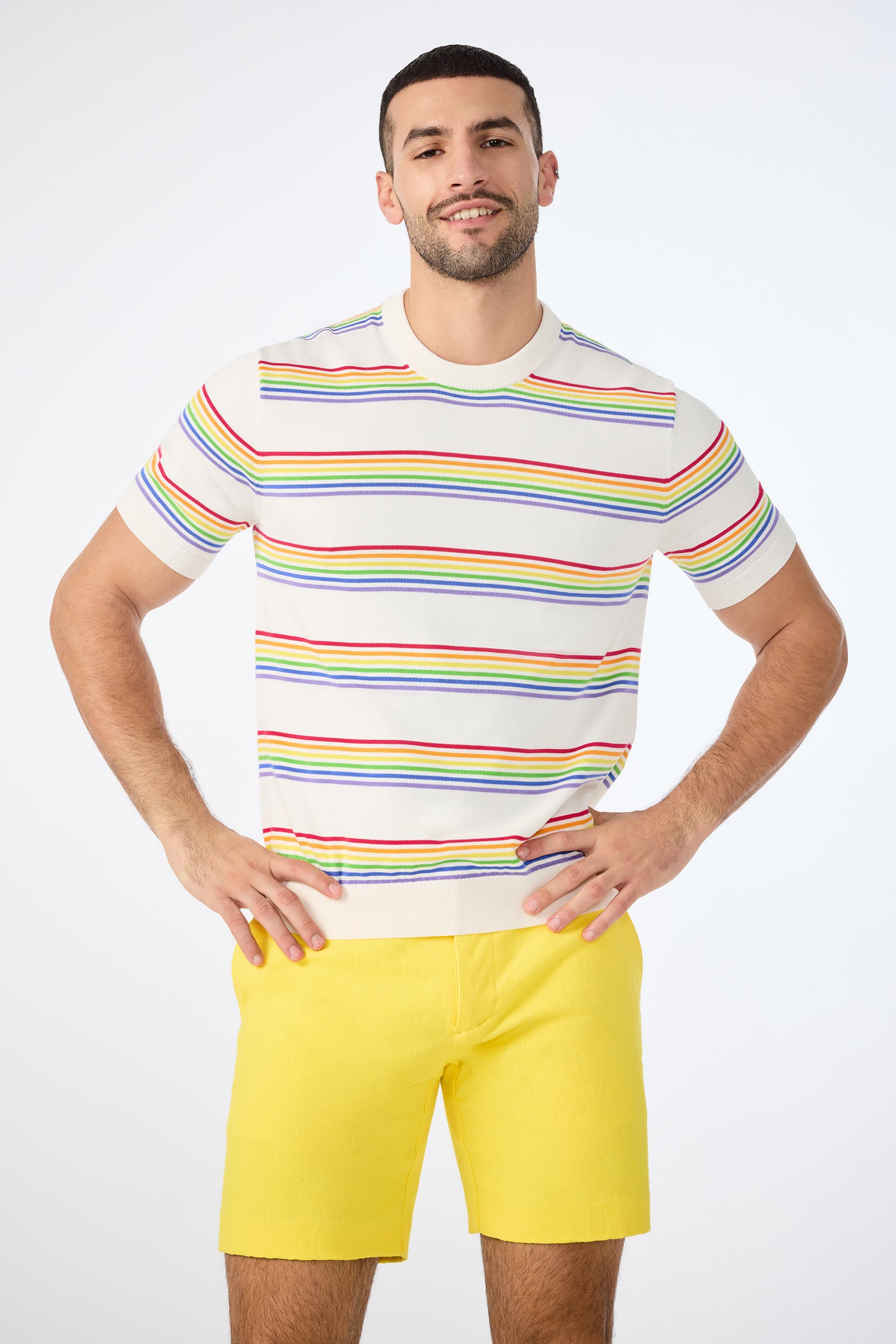 A man smiles with his hands on his hips, wearing the multi-colored Carlo Short-Sleeve Crewneck - Pride and bright yellow shorts against a plain light background.