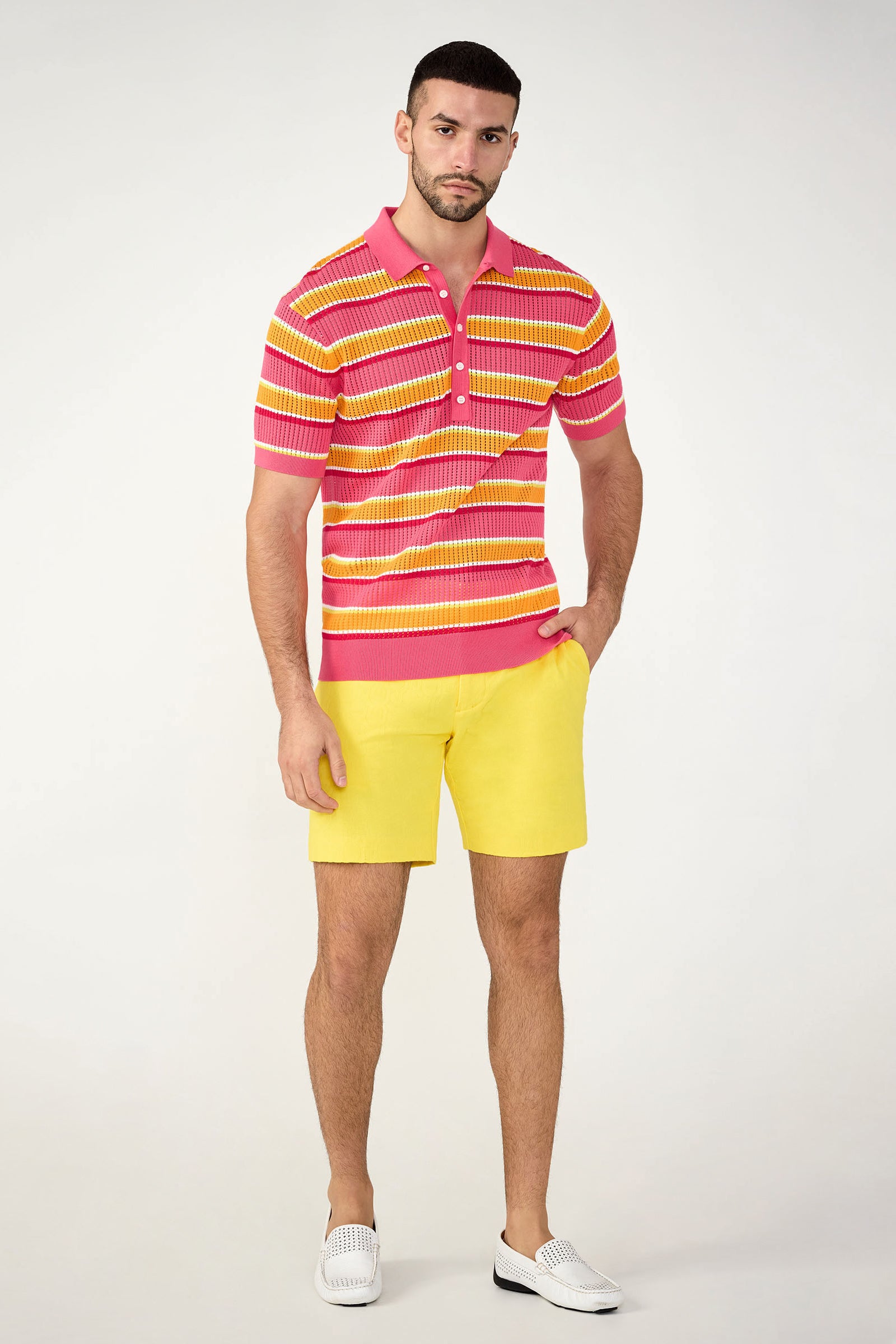 A man with short dark hair and a trimmed beard stands against a plain background, wearing the Lawrence Short - Stretch Herringbone, a pink and yellow striped polo shirt, and white slip-on shoes, with one hand in his pocket.