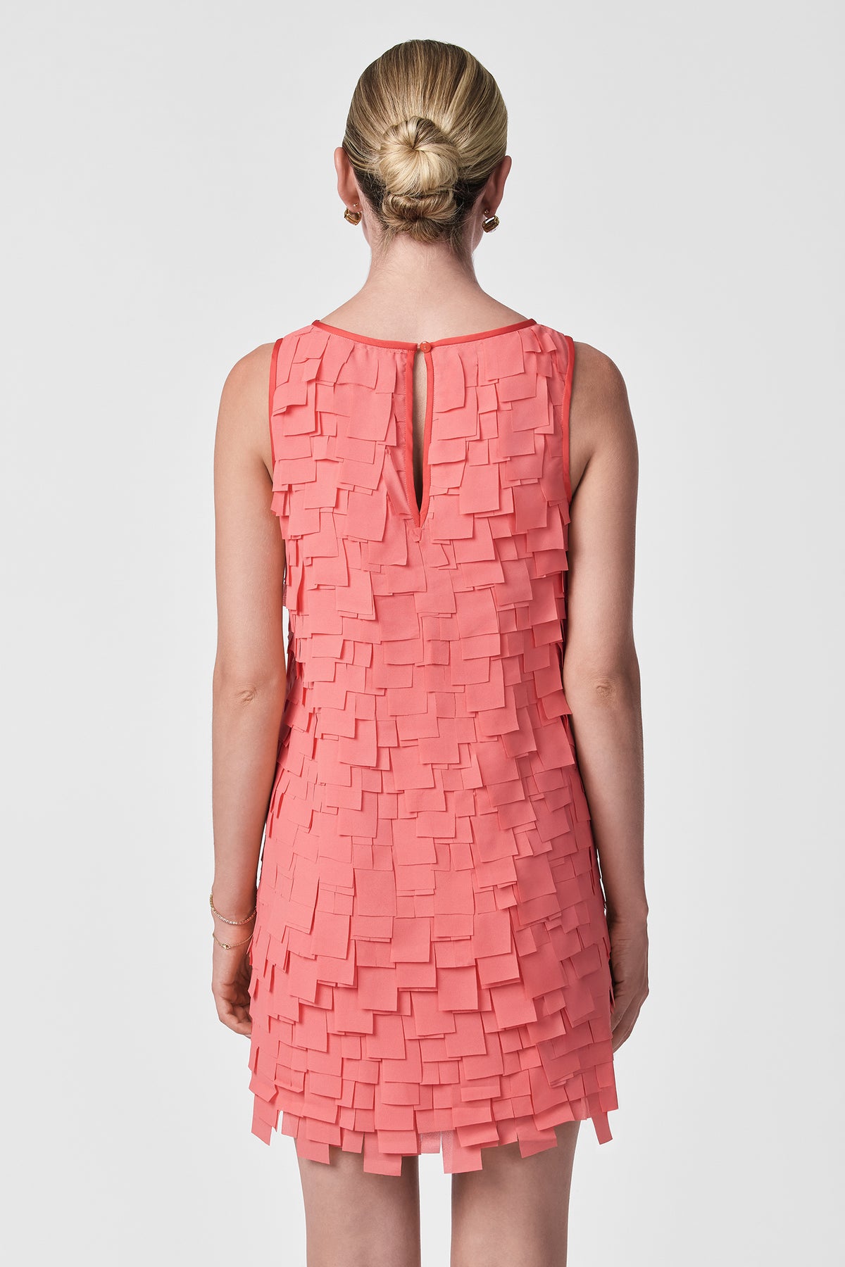 A woman with blonde hair in a neat bun is shown from the back, modeling the Valdis Sleeveless Shift Dress - Flutter Chiffon in coral, featuring layered square chiffon pieces, against a plain white background.
