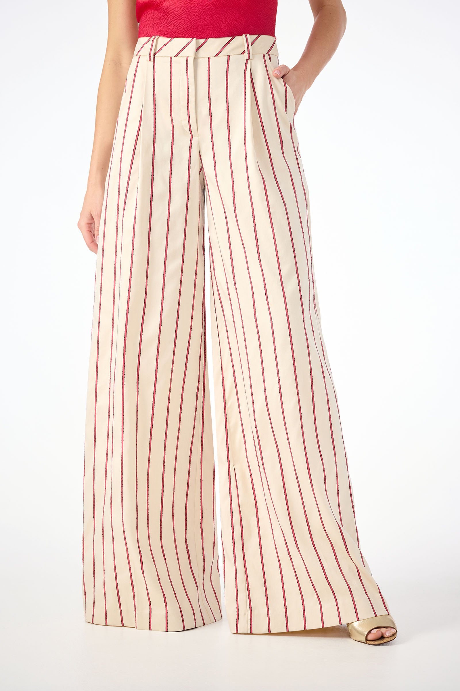 A person wearing Solani Wide-Leg Pants in Solaire Stripe, featuring high-waisted cream fabric with vertical red stripes, paired with a red top and standing against a plain white background.