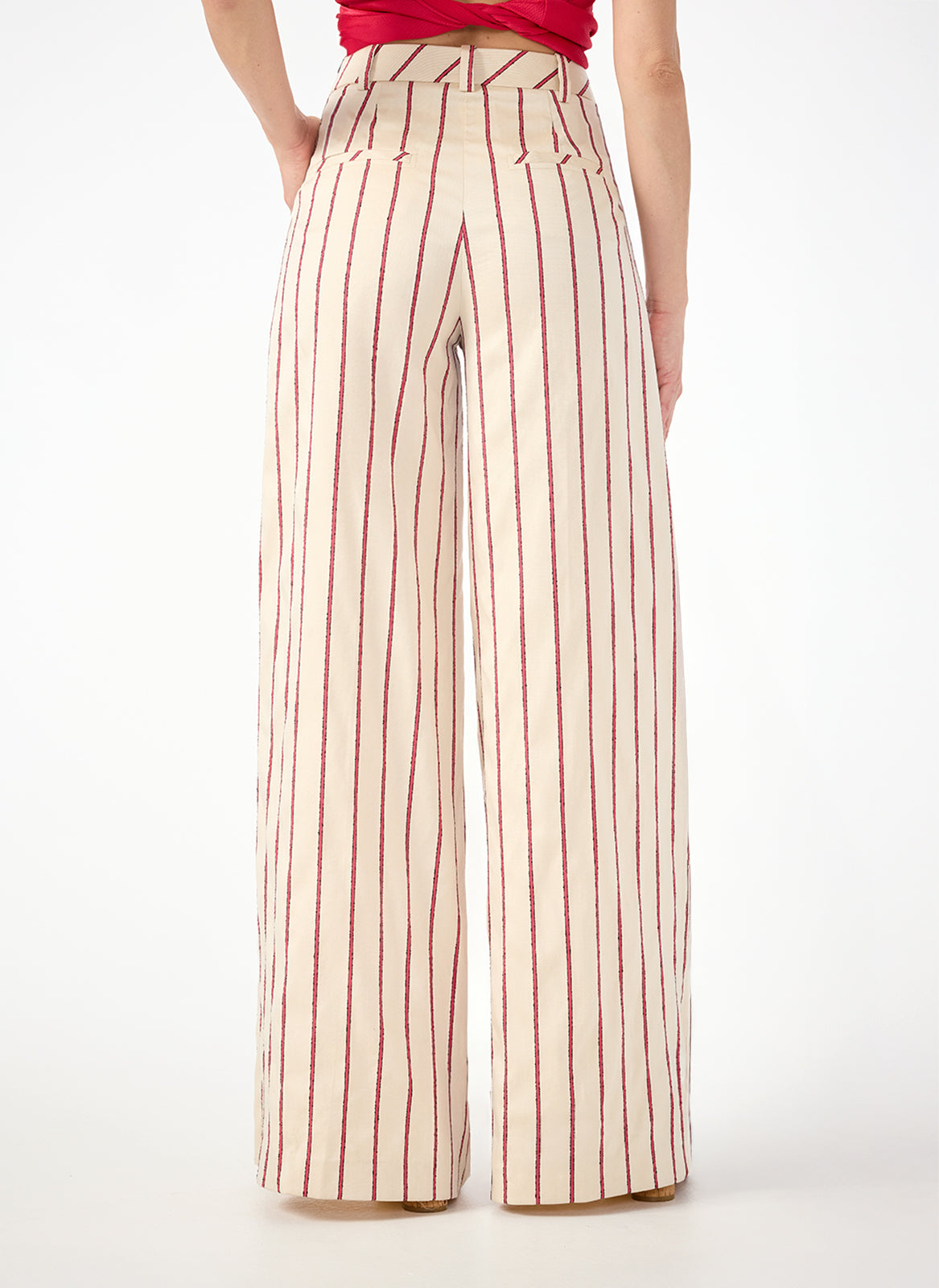 A person viewed from the back wears Solani Wide-Leg Pants - Solaire Stripe, featuring high-waisted, wide-leg cream pants with vertical red stripes and a fitted waist that flows loosely to the ankles.