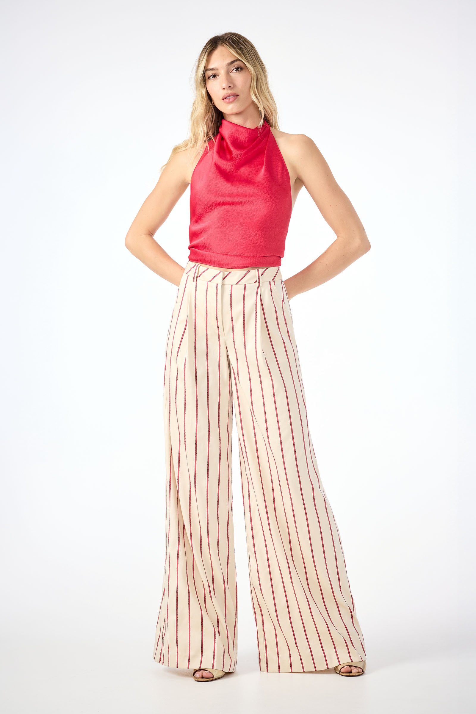 A woman faces the camera against a white background, wearing sleeveless red top and high-waisted Solani Wide-Leg Pants in Solaire Stripe. The cream pants feature vertical red stripes. Her hands are placed behind her back.