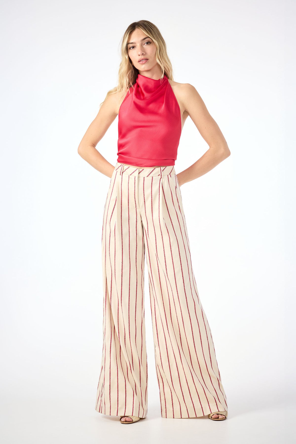 A woman faces the camera against a white background, wearing sleeveless red top and high-waisted Solani Wide-Leg Pants in Solaire Stripe. The cream pants feature vertical red stripes. Her hands are placed behind her back.