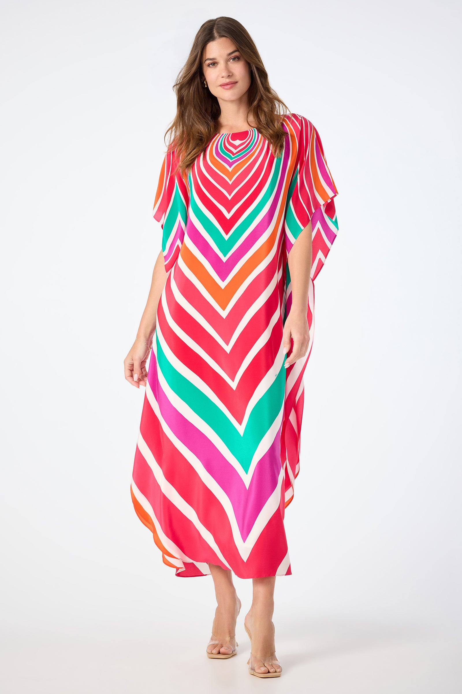 A woman in the Jalani Maxi Caftan Dress - Sur Mer, featuring bold, colorful chevron stripes, stands against a white background. She has long, wavy hair and wears clear-heeled sandals.