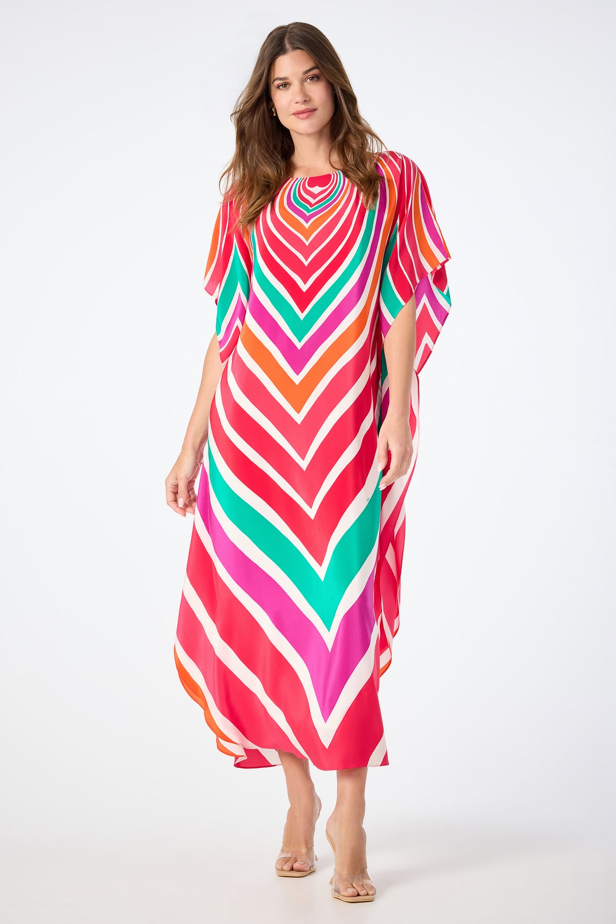A woman in the Jalani Maxi Caftan Dress - Sur Mer, featuring bold, colorful chevron stripes, stands against a white background. She has long, wavy hair and wears clear-heeled sandals.