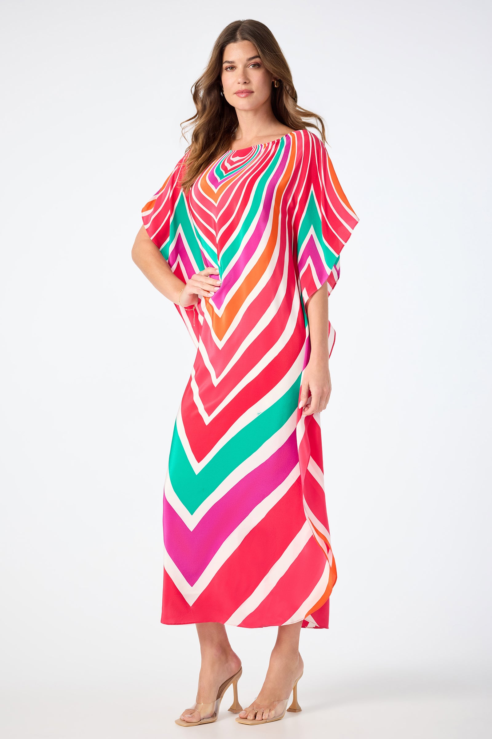 A woman with long brown hair confidently poses in the Jalani Maxi Caftan Dress - Sur Mer, featuring colorful chevron stripes in red, pink, green, and white. She pairs it with nude high-heeled sandals against a plain backdrop.