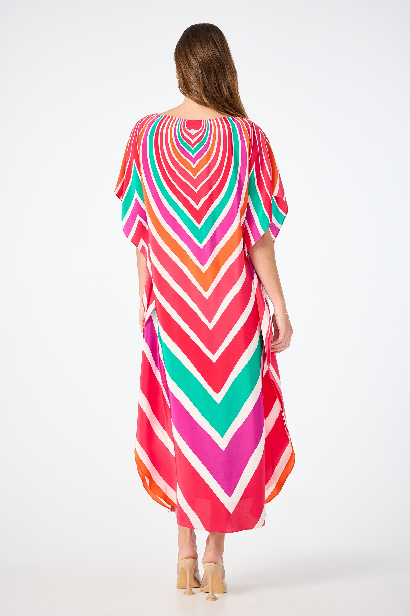 A woman with long hair, seen from behind, wears the Jalani Maxi Caftan Dress - Sur Mer featuring vibrant chevron stripes in red, pink, orange, white, and green. She pairs it with nude heels against a plain white background.