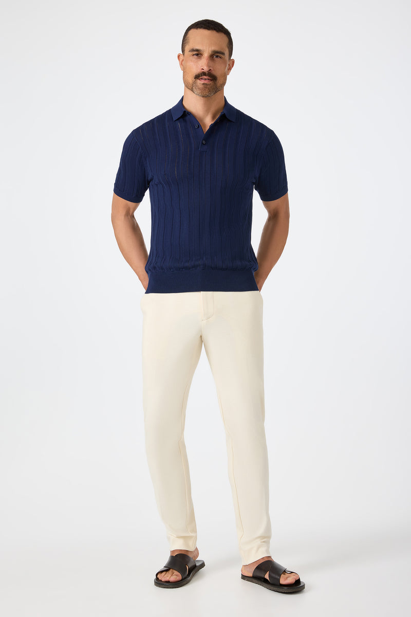 A man with short hair and a mustache stands against a white background, wearing cream-colored Alex Slim Trouser - Ecru, a navy knit polo shirt, and black sandals with his hands in his pockets.