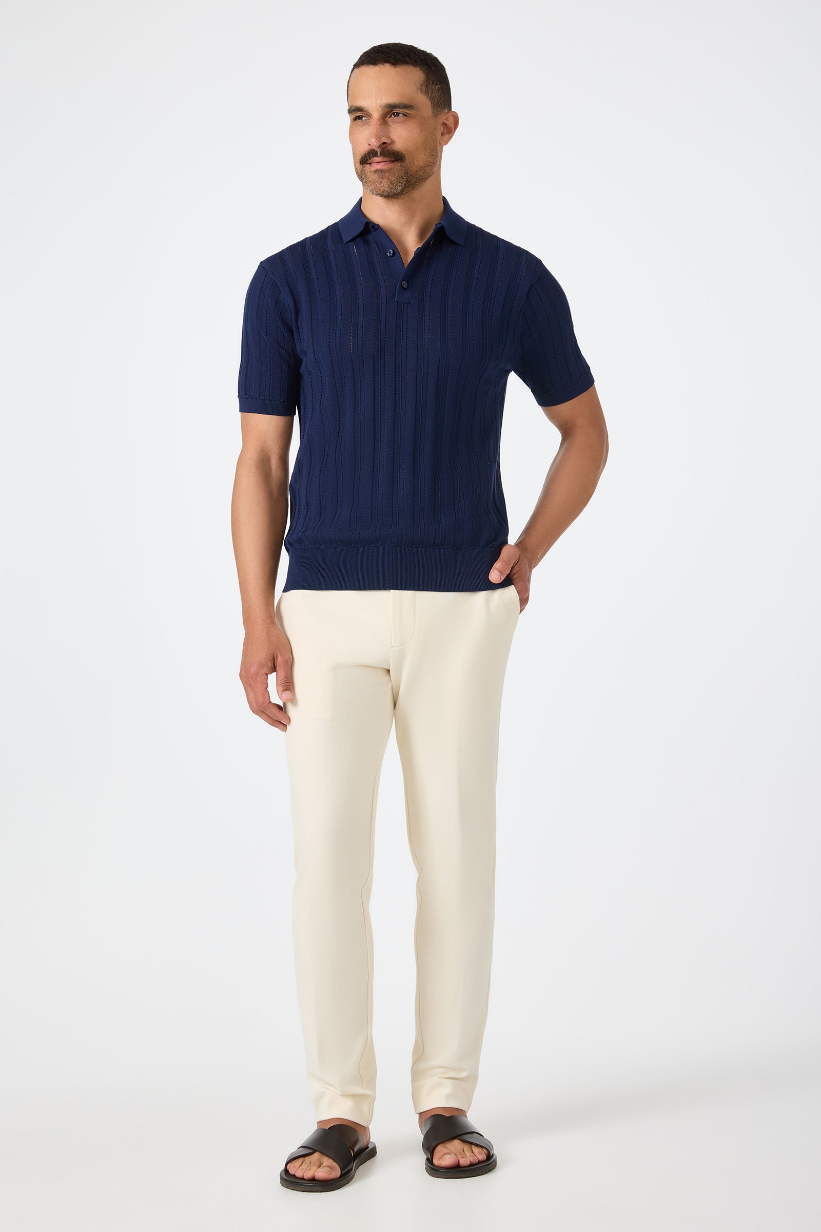 A man stands against a plain background wearing the Kensington Short-Sleeve Polo in navy blue, cream-colored pants, and black sandals, with one hand in his pocket.