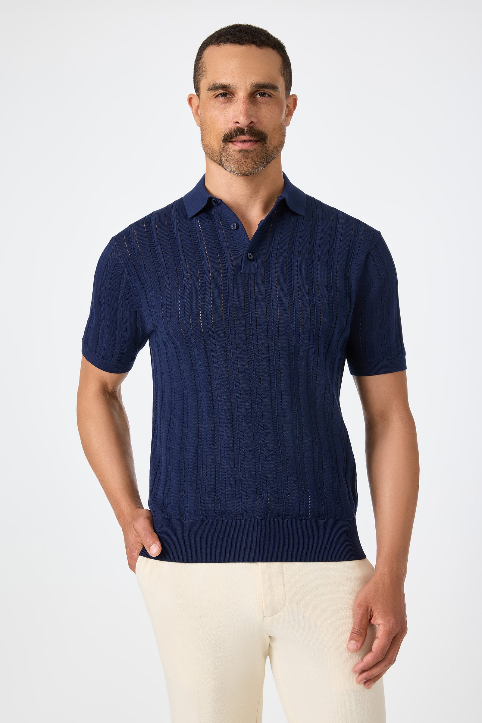 A man with short hair and a mustache wears the Kensington Short-Sleeve Polo in Ink with light beige pants, standing against a plain white background with one hand in his pocket.