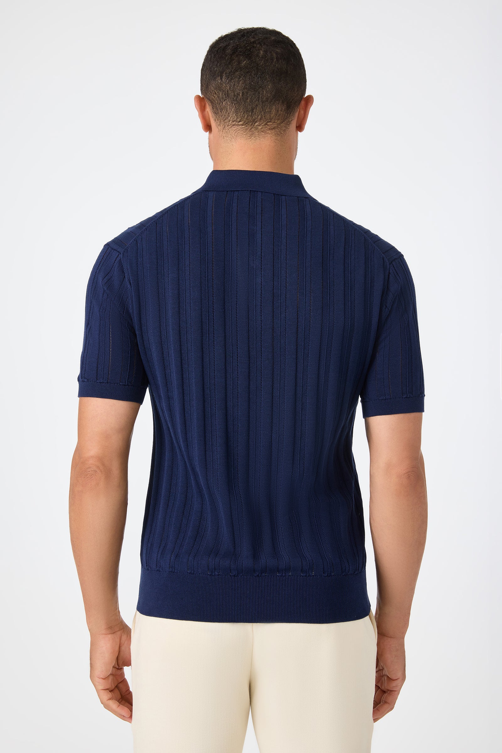 A man stands with his back to the camera, wearing the Kensington Short-Sleeve Polo in navy blue and cream-colored pants against a plain white background.