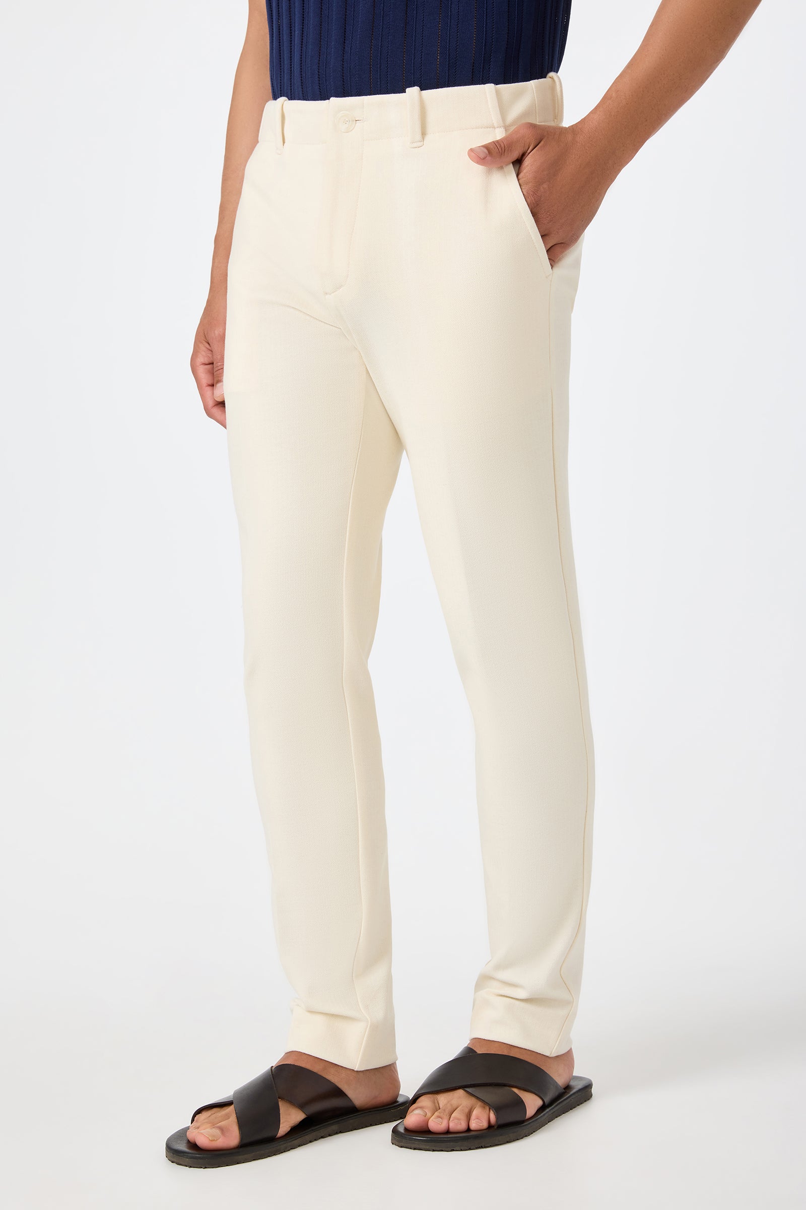 Wearing the Alex Slim Trouser in Ecru with dark brown sandals and a navy textured shirt, the person stands with one hand in their pocket against a plain white background.