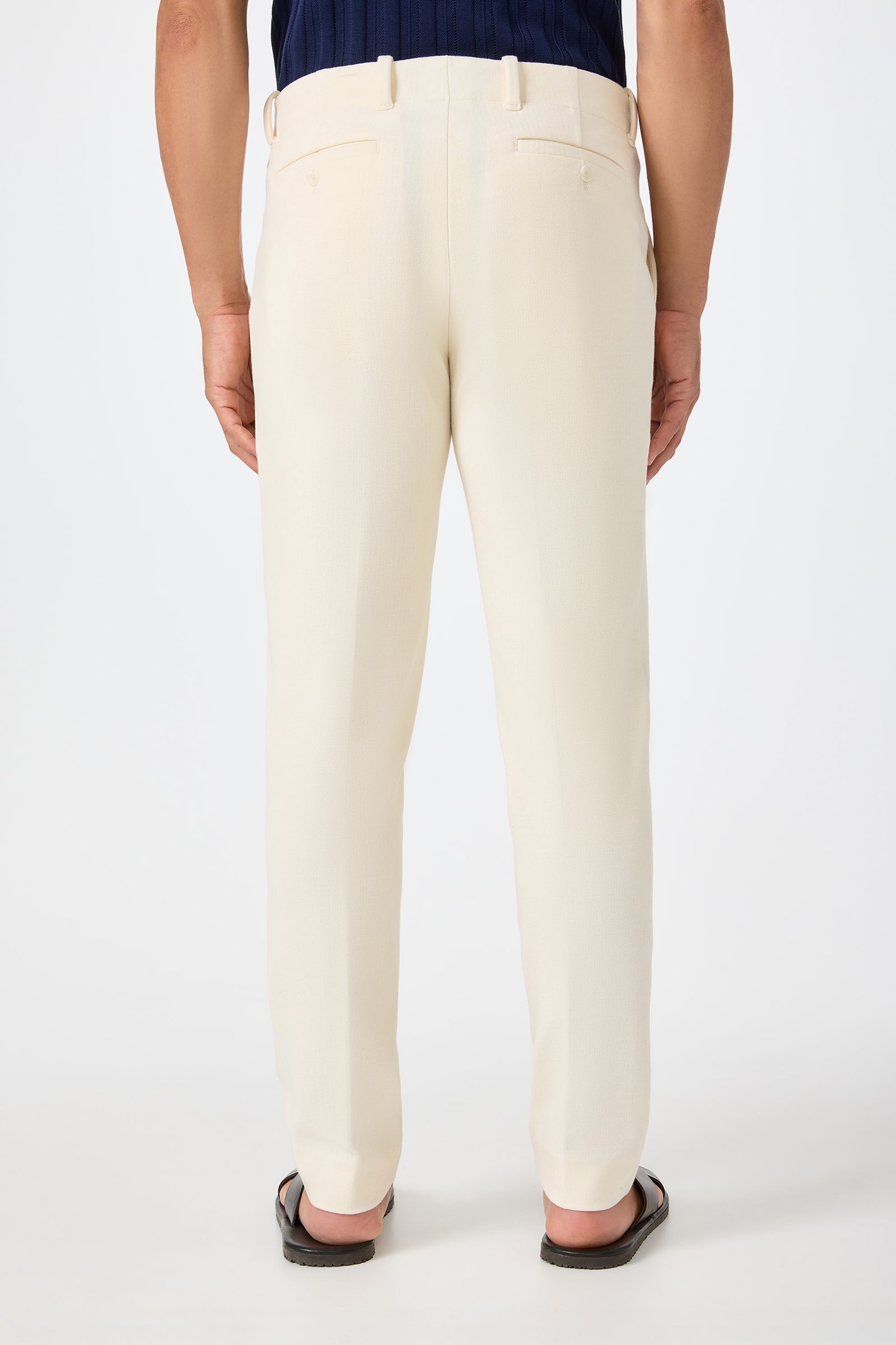 A person is seen from behind wearing the Alex Slim Trouser in Ecru with a dark shirt and black sandals, standing against a plain white background.
