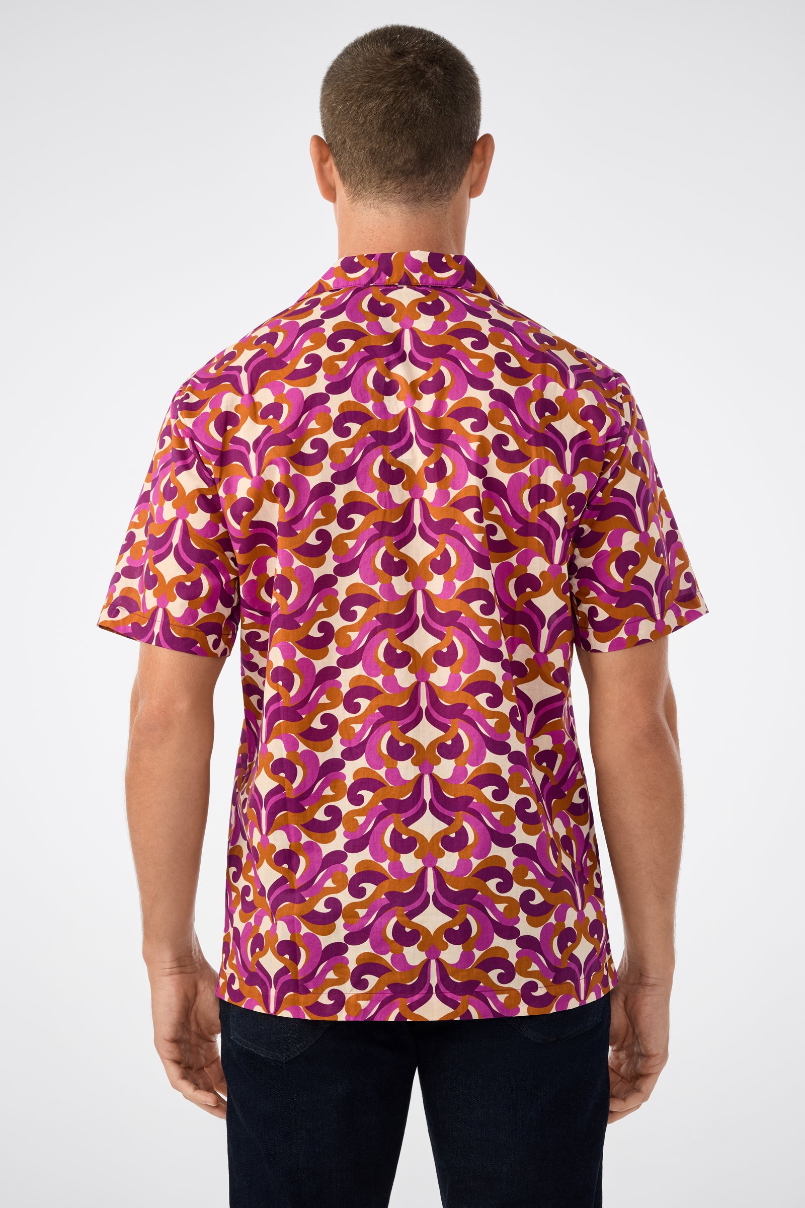 A man faces away, wearing the Felix Ss Camp Shirt with a bold purple, pink, yellow, and white geometric pattern, paired with dark pants against a plain light background.