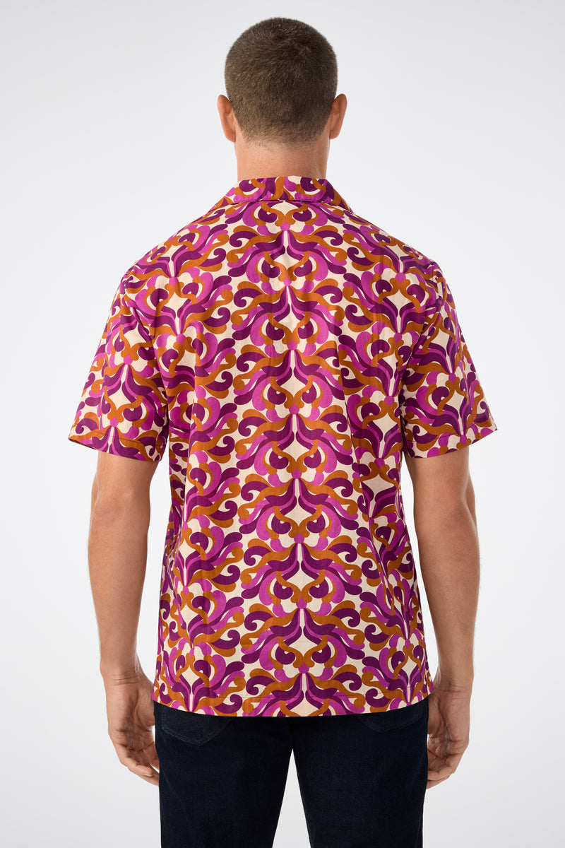 A man faces away, wearing the Felix Ss Camp Shirt with a bold purple, pink, yellow, and white geometric pattern, paired with dark pants against a plain light background.